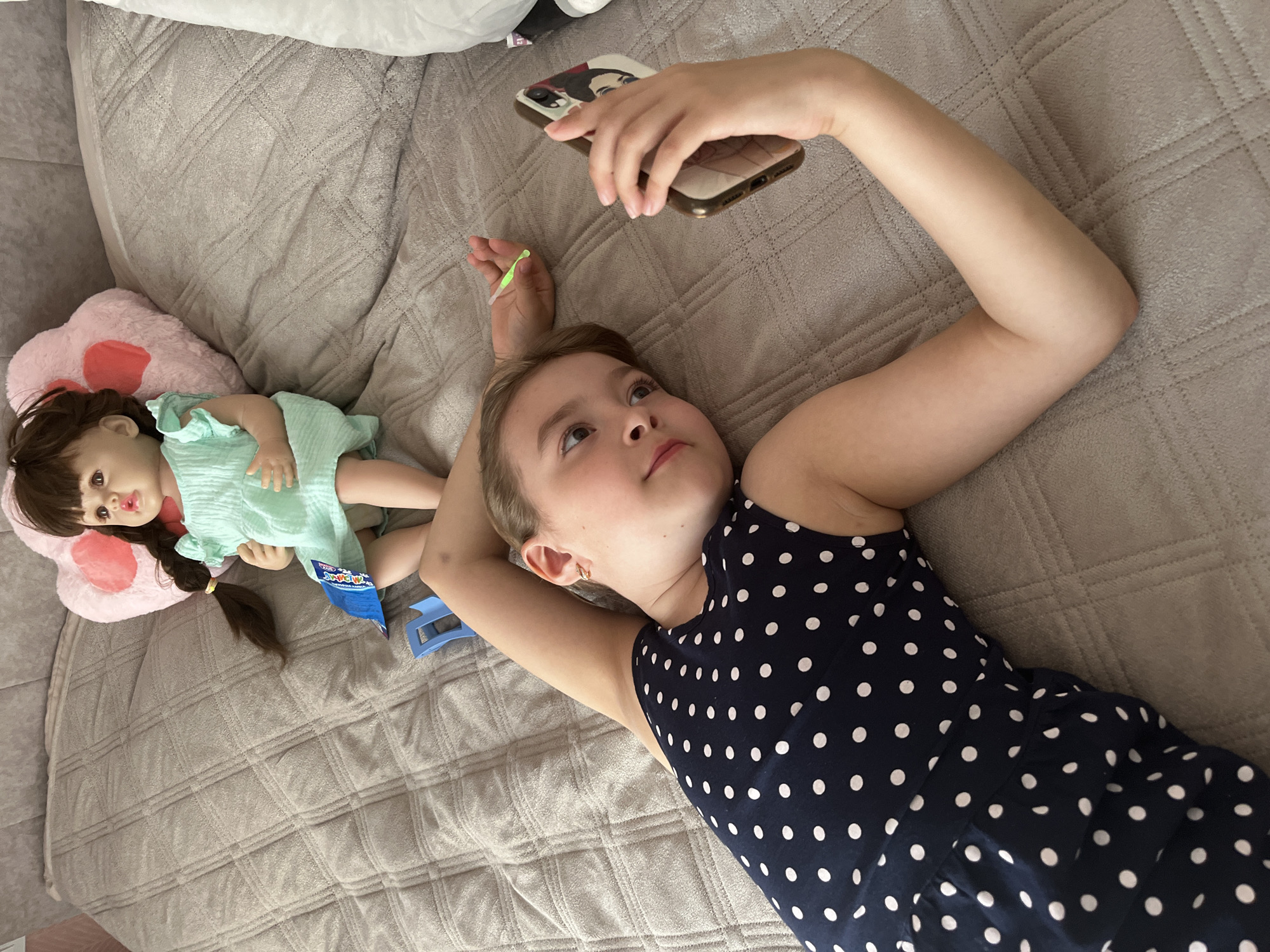 A girl is lying on a gray quilted bed, holding a cell phone in her right hand, looking up and to the left. She is wearing a navy blue dress with white polka dots. A doll in a green dress is lying on a pink heart-shaped pillow to the left of the girl. Next to the doll is a blue plastic object and a blue package. The image was likely taken indoors. The location mentioned is Новая Чернушка, Russia.