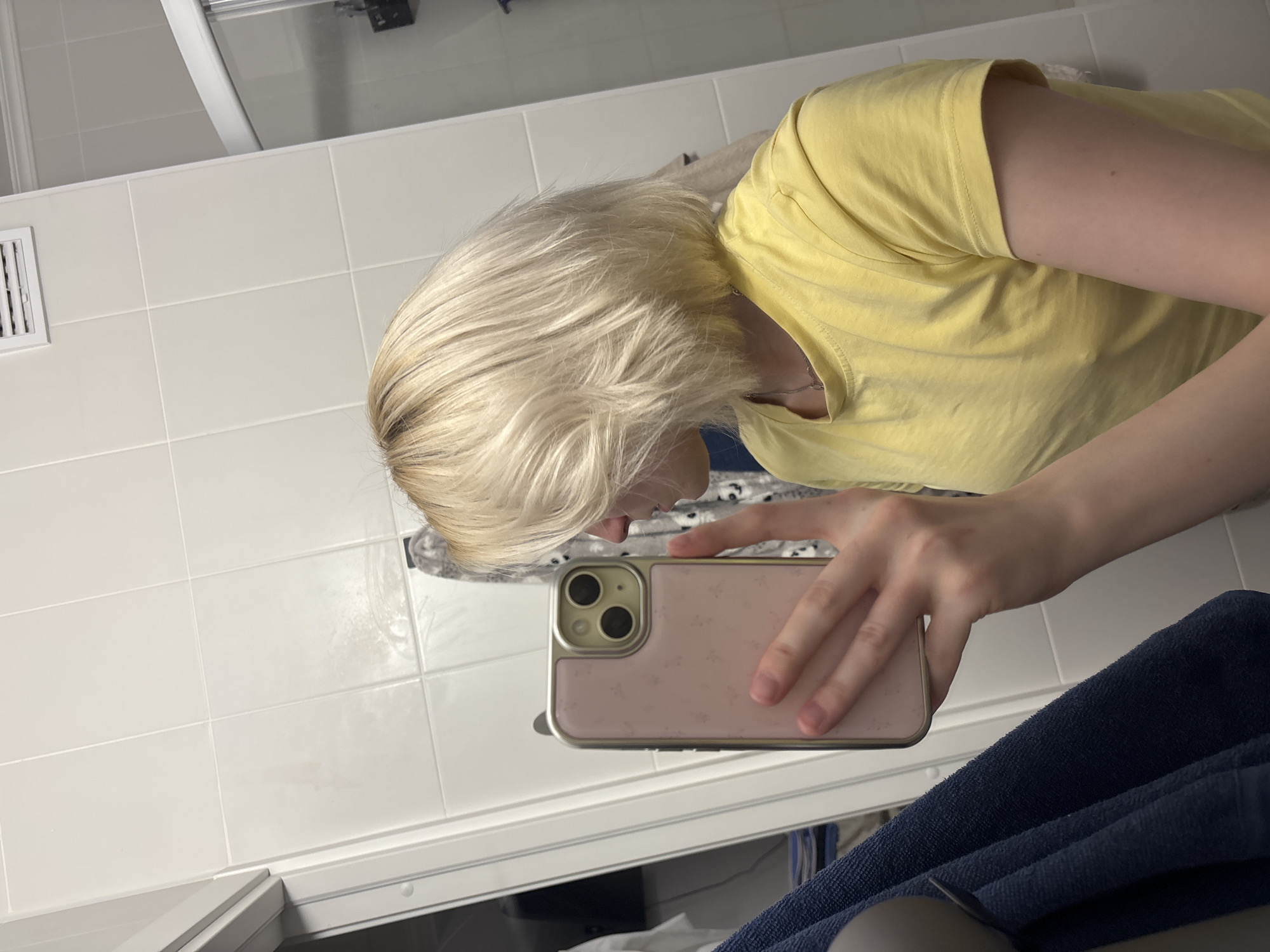 A person with blonde hair wearing a yellow t-shirt is holding a pink cell phone and taking a picture in a bathroom. The bathroom has white tile walls and a blue towel is draped over the edge of the sink. A vent is visible on the wall. The location is Unknown, Unknown.