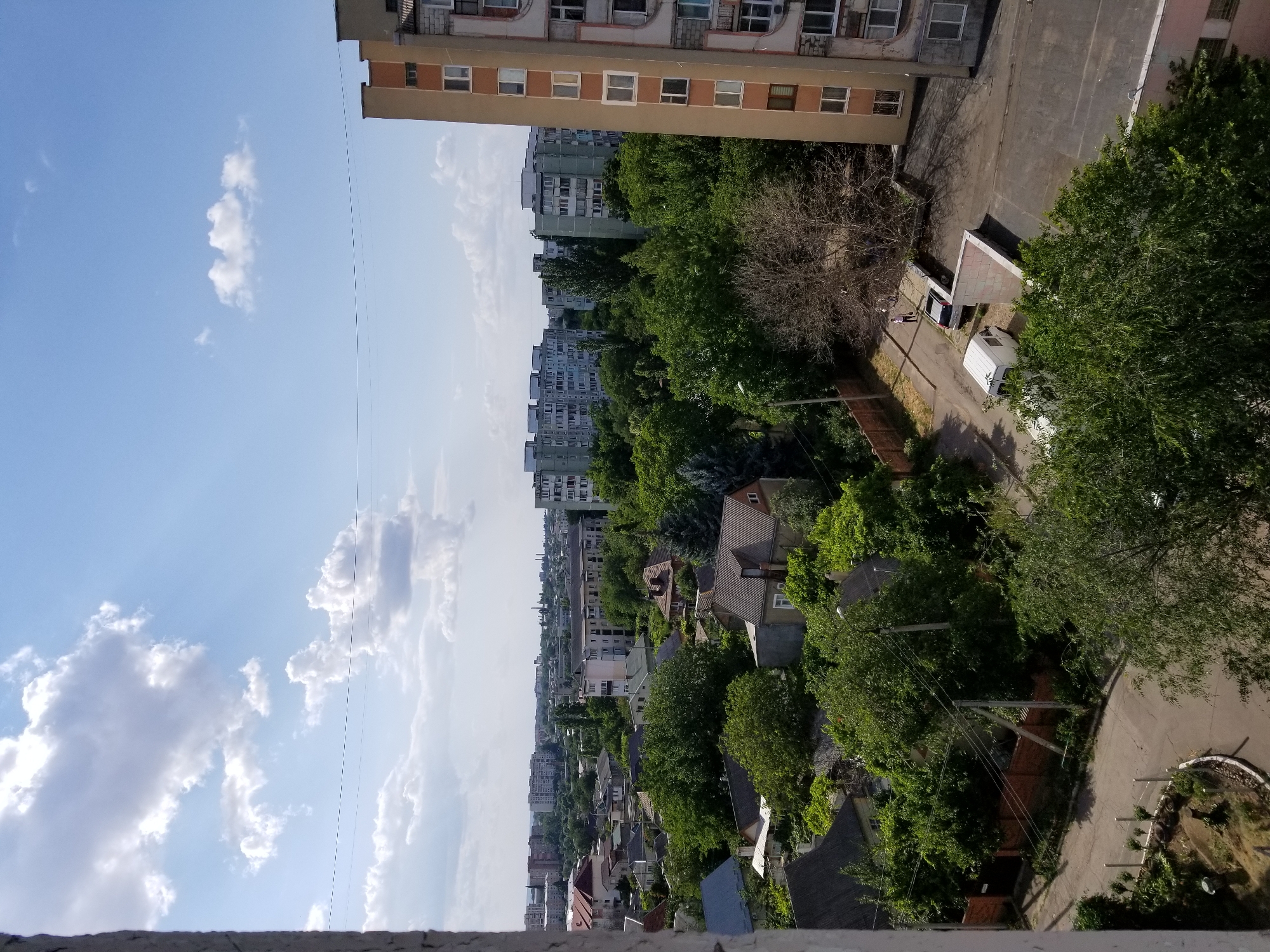 An aerial, slightly angled shot captures a section of Tiraspol, Moldova, on a partly cloudy day. The foreground is dominated by a concrete structure on the left, resembling a building corner, obscuring part of the view. Beyond this, a dense canopy of green trees stretches across the mid-ground, interspersed with the roofs of various houses and buildings. Red-tiled roofs are visible on several houses, alongside some with grey shingles. In the distance, numerous apartment blocks, characteristic of Soviet-era architecture, punctuate the skyline, with their multi-story facades and numerous windows. A tall, multi-story building with a beige and orange facade stands prominently on the right side of the frame. Streets are visible winding through the residential areas, with a few vehicles, including a white van and a car, parked or moving. A lone figure, appearing to be a person, is walking on the pavement near the beige and orange building. The sky above is a bright blue, with scattered white clouds casting shadows on the landscape below. Power lines are visible crisscrossing the upper portion of the image.