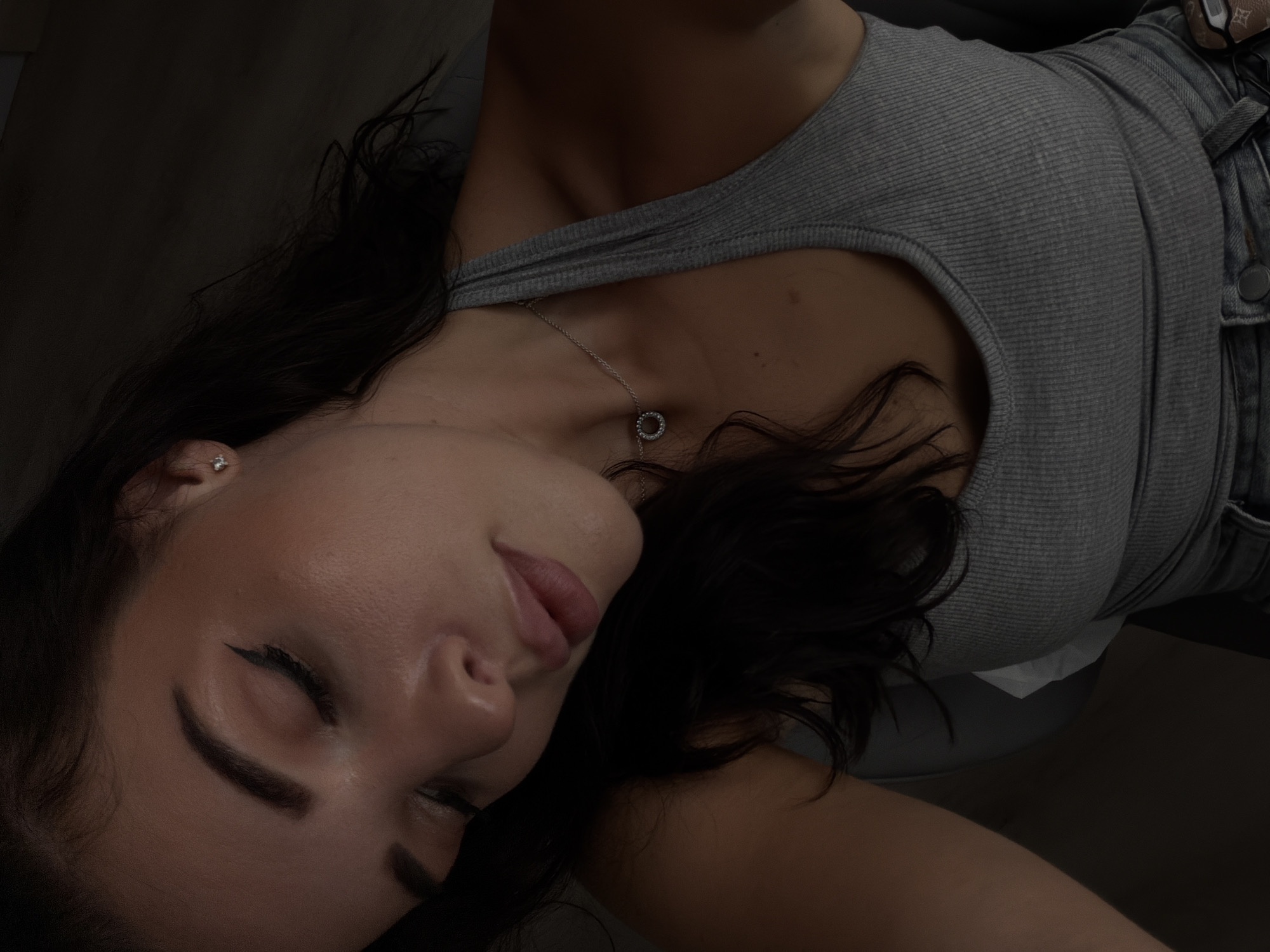 The image shows a woman in a grey ribbed tank top and blue jeans. She has long dark hair, closed eyes, and defined eyeliner. She wears a silver necklace with a circular pendant and a stud earring. Part of a brown designer purse is visible in the top right corner. A white tissue is partially visible beneath her tank top. The setting is dark and dimly lit. There is no indication of the location being Bac Nha Trang Ward, Vietnam.
