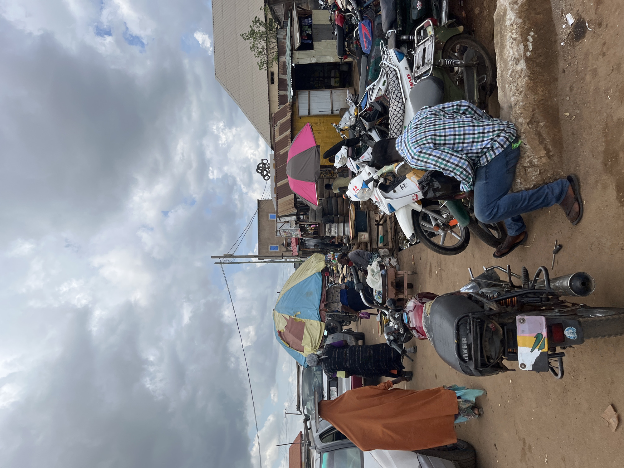 The image depicts a busy outdoor market scene in Rafin Par, Nigeria, under a cloudy sky. Several motorcycles are parked or being worked on, suggesting a transportation hub or repair area within the market. People are seen interacting with the motorcycles and shopping.
In the foreground, a person wearing a blue and green checkered shirt and jeans is crouched down, appearing to work on a motorcycle. To the right, a large boulder or concrete structure lines the edge of the scene. Further back, a collection of tires and more motorcycles are visible. Several colorful umbrellas, one pink and gray and another with yellow and blue sections, provide shade over market stalls.
People in traditional attire are also present, with one person in an orange wrap and another in a dark patterned dress. The architecture of the buildings in the background appears simple, with corrugated metal roofs and makeshift storefronts. The overall impression is one of an active and bustling local marketplace. The presence of many motorcycles and the repair activity hints at a place where these vehicles are essential for transportation and commerce.
