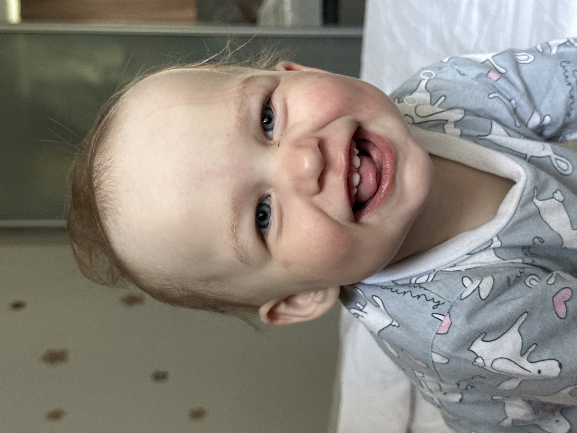 A close-up shot depicts a baby with light skin and blue eyes, wearing a gray pajama set patterned with white bunnies. The baby is smiling broadly, revealing a few teeth and their tongue, mouth open. The background appears to be indoors with a blurry wall with brown and green spots. The location or context of the photo in the city of Chaikovsky, Russia, cannot be determined from the image.