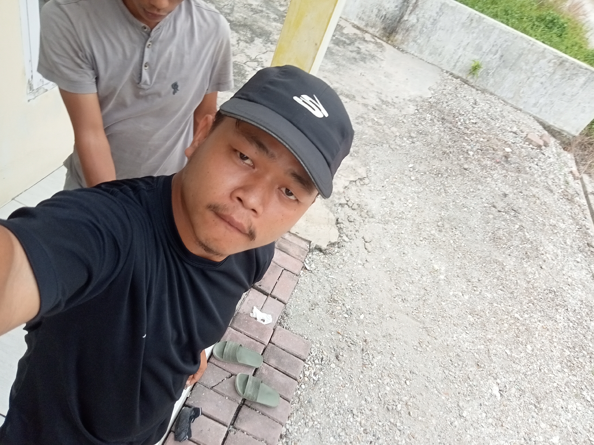 In the city of Kualu, Indonesia, two individuals are captured in a selfie-style photograph. The foreground features a man wearing a black t-shirt and a black baseball cap with a white logo. He is positioned in a way that his face and upper body are prominently displayed, with his left arm extending towards the camera. To his right, a pair of green slide sandals are placed on a brick pathway. A white tissue is also visible on the pathway near the sandals. In the background, another person wearing a grey t-shirt is partially visible, with their head and upper torso in focus. They appear to be standing behind and slightly to the left of the person in the foreground. The environment consists of an outdoor setting with a concrete ground, gravel, and a low concrete wall with some vegetation. A yellow vertical pillar is also present in the background, suggesting an urban or semi-urban location. The lighting suggests it is daytime.
