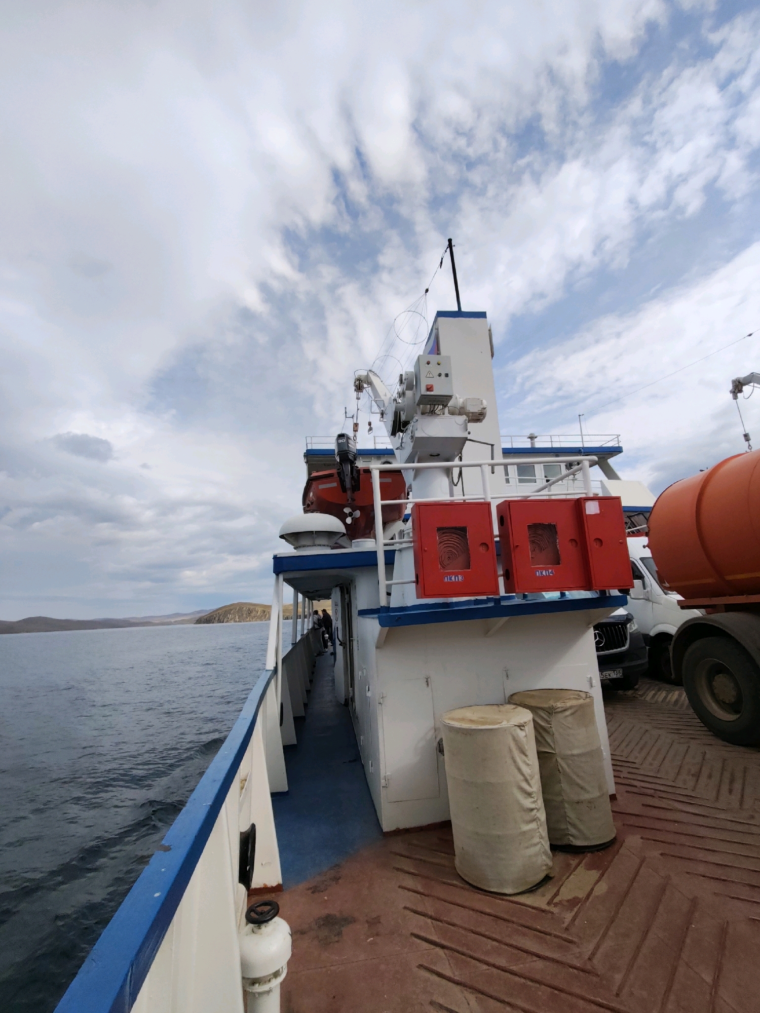 The image captures a view from the deck of a ferry, looking towards the stern and right side. The ferry is at sea, with a calm, choppy blue water visible to the left. In the distance, a hilly, treeless coastline is discernible under a cloudy sky. On the deck of the ferry, a white structure with blue trim is prominent. This structure houses a red lifeboat, two red fire hose cabinets labeled with Cyrillic text, and various control panels and equipment mounted on a mast. Two cylindrical objects covered in beige fabric are positioned on the deck in front of the white structure. To the right, part of an orange tanker truck is visible, along with its large rear wheel. Behind the tanker truck and white structure, a white van is parked. A few individuals are standing near the railing of the ferry on the left side of the image, observing the scenery. The deck itself is made of reddish-brown metal with a patterned surface. The overall impression is one of a working ferry transporting vehicles and people across a body of water, likely on a cloudy day. The location is the city of Sakhiurta, Russia.
