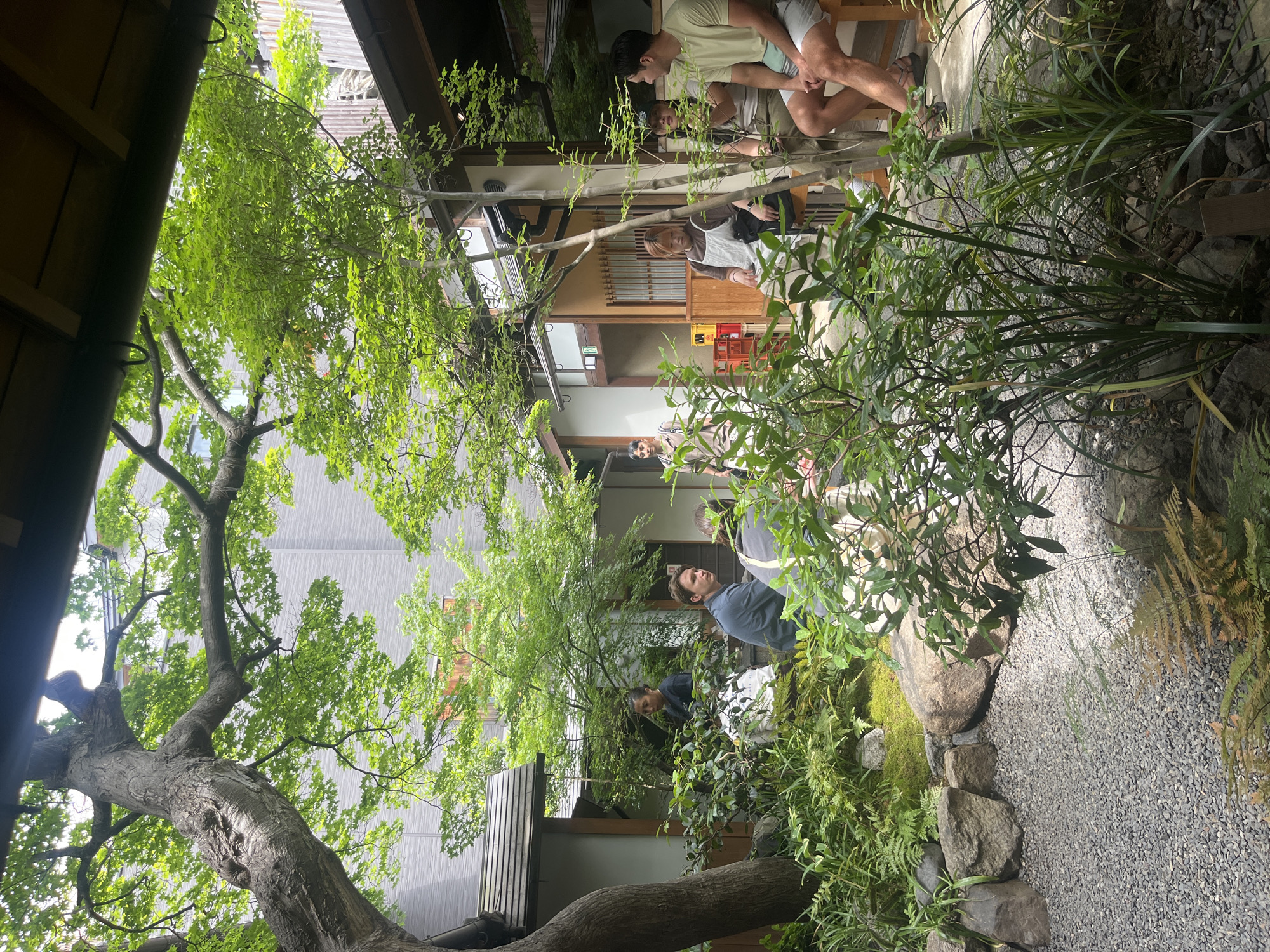 The image captures an outdoor courtyard setting in Kyoto, Japan. The scene is a mixture of traditional Japanese architecture and lush greenery. People are present in the courtyard, engaged in relaxed activities such as sitting and standing. The architecture features wooden structures, sliding doors, and tiled roofs. The greenery consists of trees, shrubs, and moss-covered rocks. A gravel path leads through the courtyard. The overall environment suggests a peaceful and tranquil atmosphere.