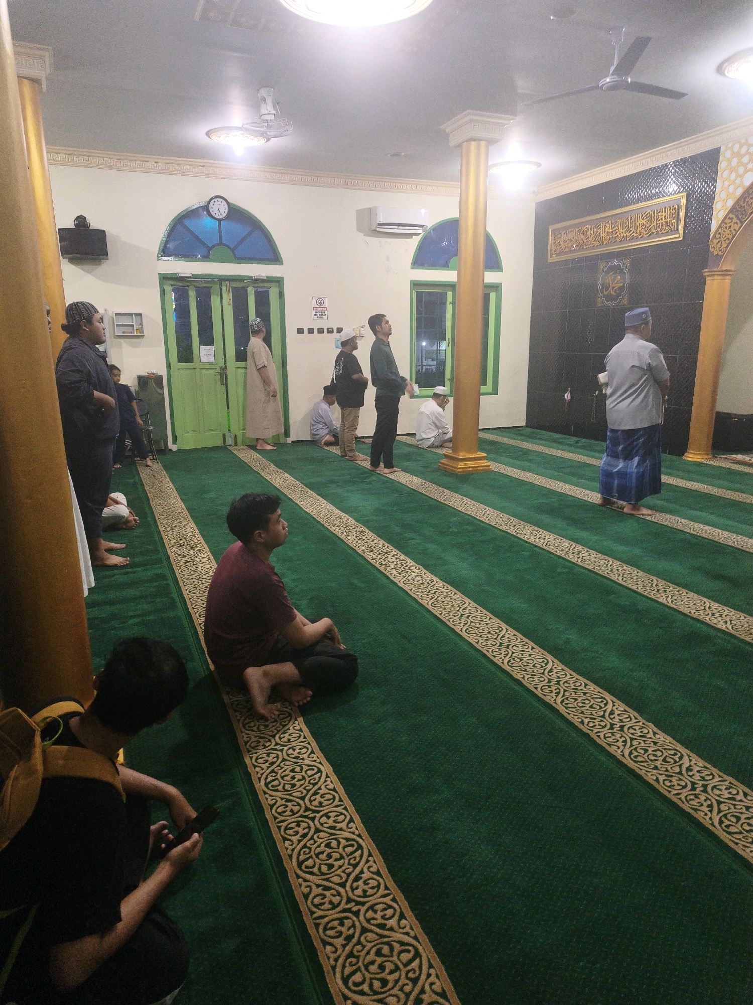 The image depicts the interior of a mosque in Samarinda, Indonesia. The mosque features green carpeting with decorative gold borders. Several men are present, some standing in prayer rows, while others are seated on the floor. In the foreground, two men are visible; one is seated and looking up, and another is in a squatting position, holding a mobile phone. The background reveals a large clock on the wall, green double doors, and an air conditioning unit. A decorative frame with Arabic calligraphy is mounted on the wall. The overall scene suggests a quiet and reverent atmosphere during a religious gathering.
