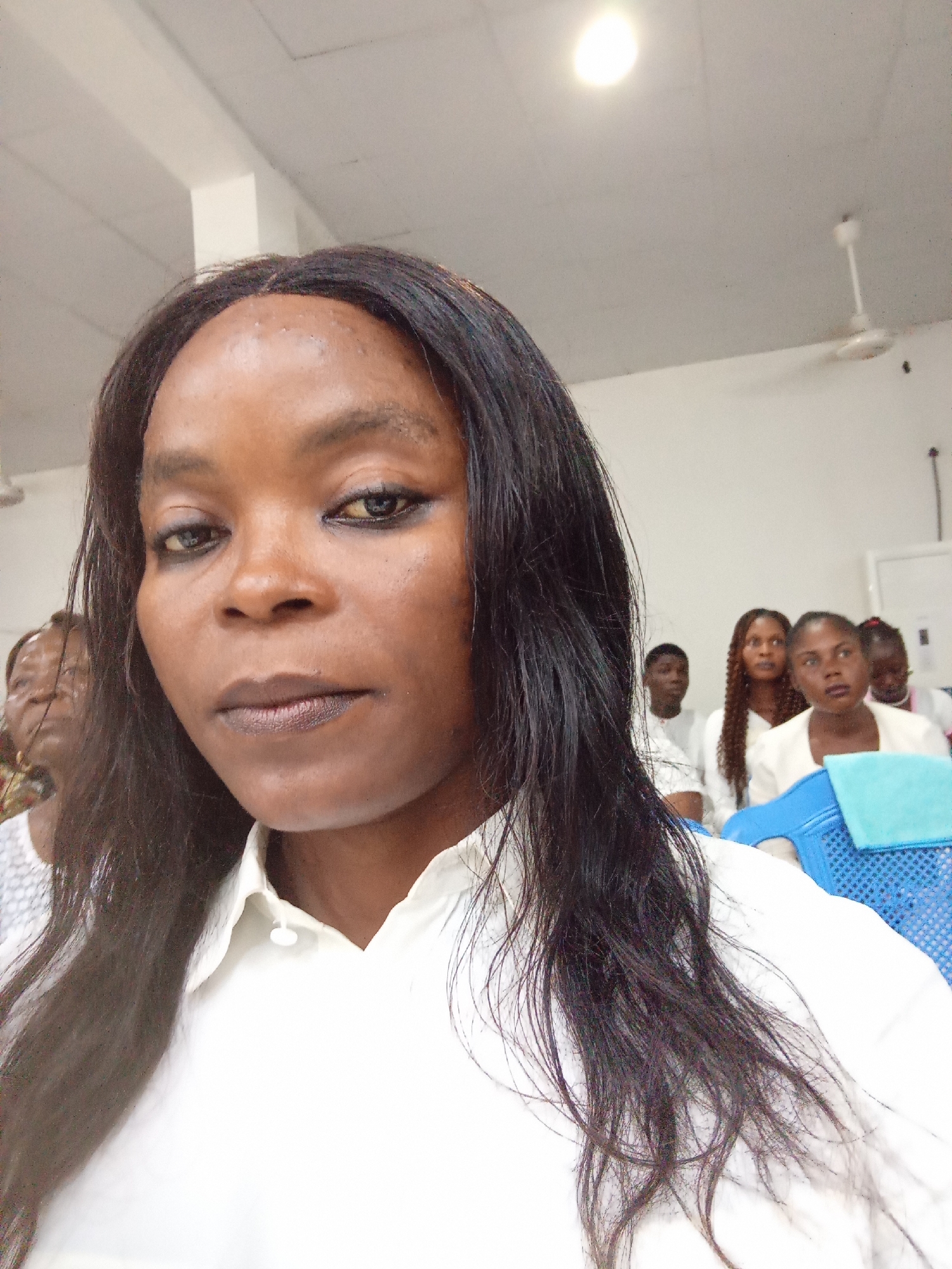 A woman with long, dark hair and dark makeup is taking a selfie in an indoor setting. She is wearing a white collared shirt. Behind her, several other people are seated, all appearing to be wearing white clothing. The background suggests an event or gathering, possibly in a community hall or place of worship. The walls are white, and there are ceiling fans and fluorescent lighting visible. A blue plastic chair with a light blue towel draped over it is in the foreground to the right. The scene appears to be well-lit.