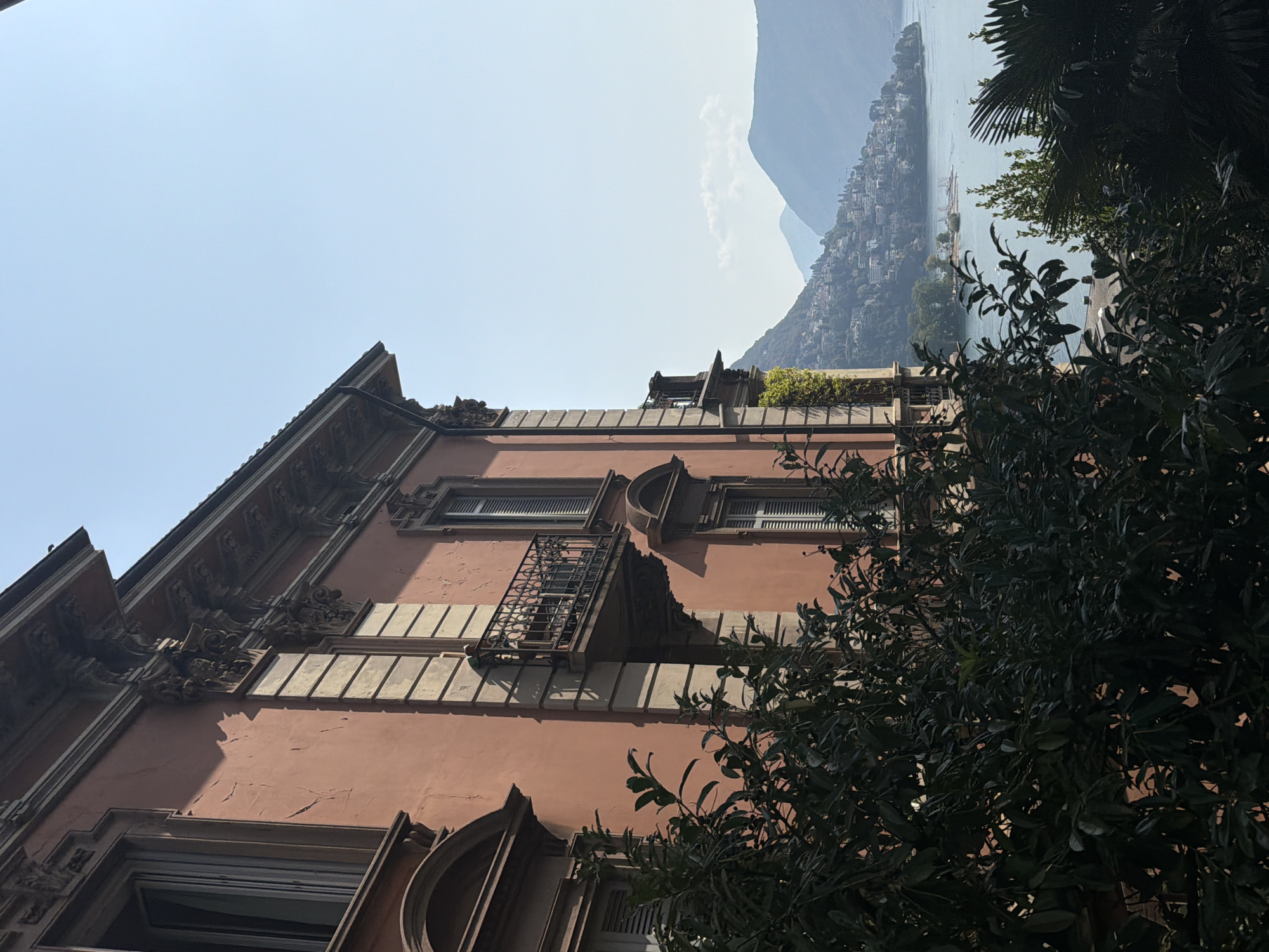 A side view of a salmon-colored building with architectural details is shown in the foreground, partially obscured by lush green foliage. The building features a balcony with decorative ironwork on the second floor, and ornate cornices above the windows. Beyond the building, a body of water, likely Lake Lugano, stretches out towards rolling hills covered in buildings and trees, indicating the city of Lugano, Switzerland. The sky is a clear, pale blue. The scene appears to be captured on a bright, sunny day.