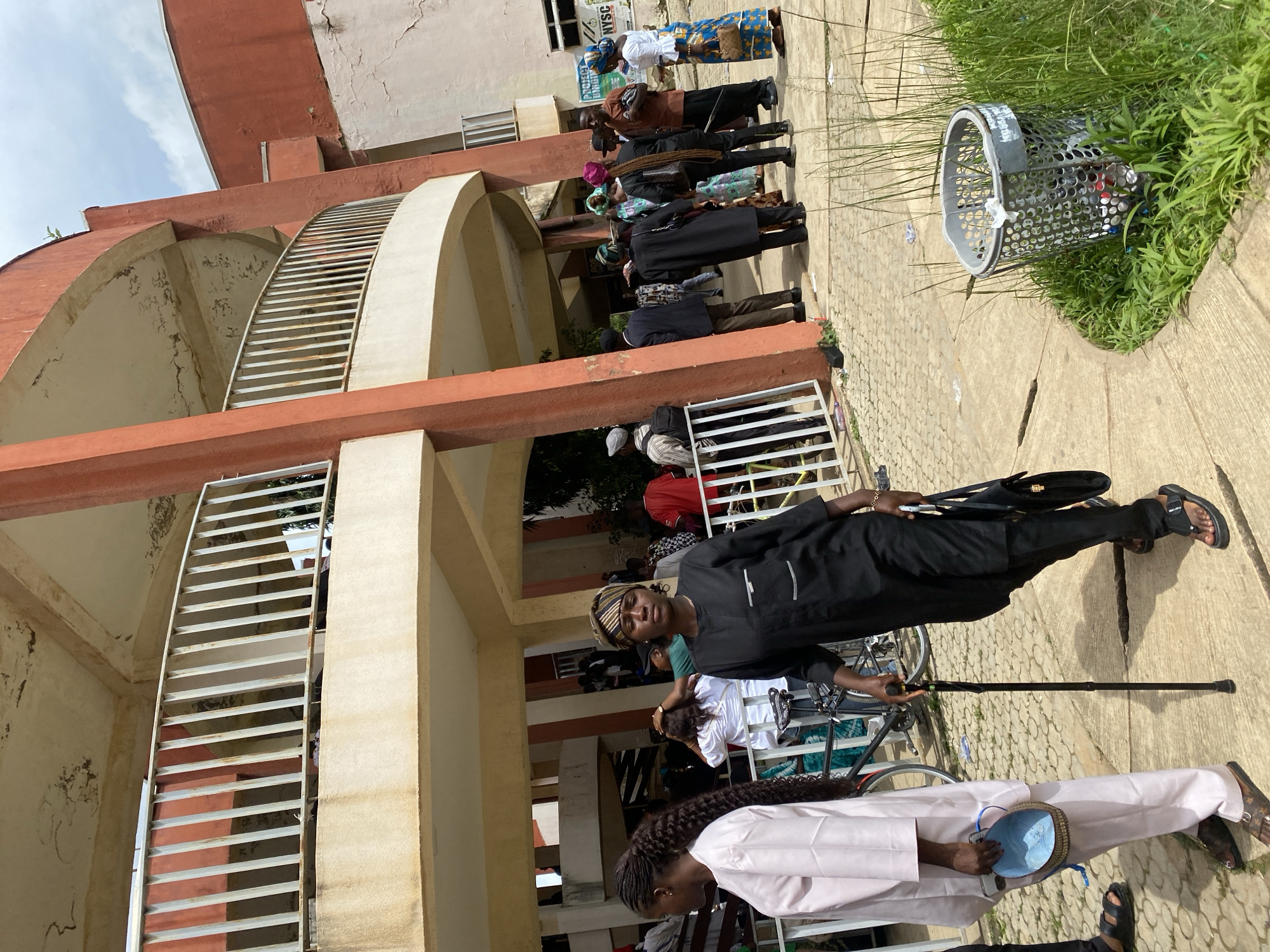 In Jos, Nigeria, an outdoor scene unfolds with a group of people gathered near a reddish-orange, multi-story building. The building features a distinctive curved balcony with white railings, showing signs of wear and tear on its concrete surfaces. People, dressed in various traditional and casual attire, are standing and milling about on a paved walkway. Some individuals are holding bags, and one person is using a cane for support. A bicycle is visible among the crowd. In the foreground, a wire mesh trash can filled with debris sits amidst lush green grass. The environment appears to be a public space, possibly associated with an institution, given the presence of a sign that reads "PROJECT BINDING" and another possibly indicating "NYSC." The sky is partly cloudy.
