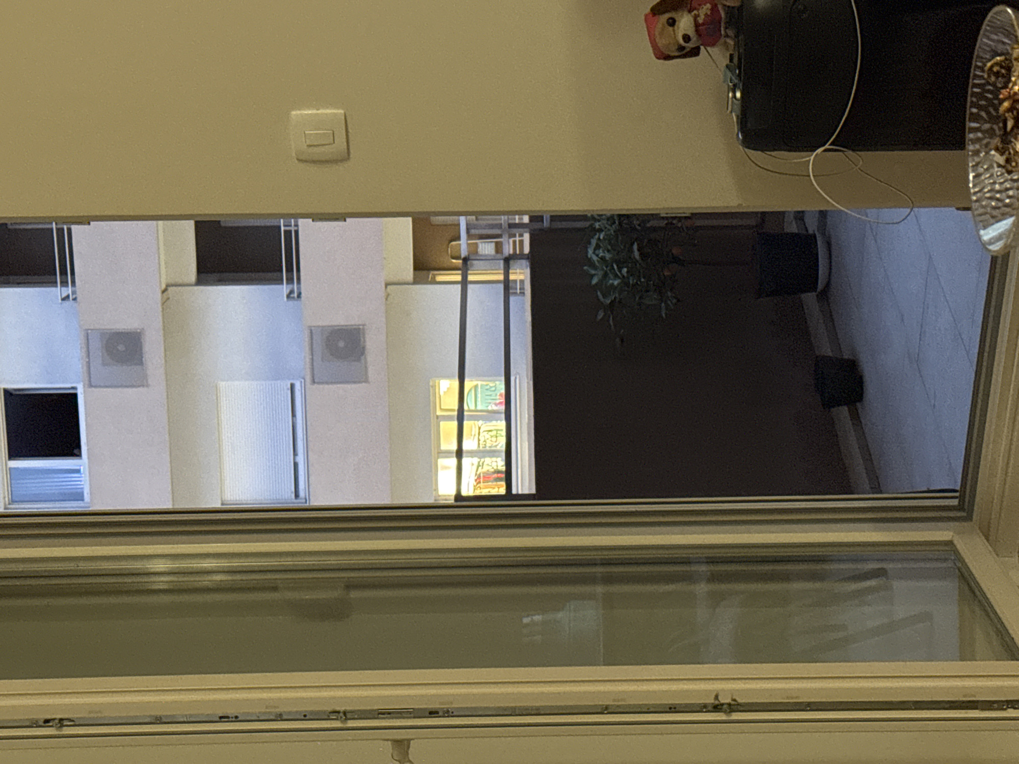 The image depicts a scene viewed through an open doorway or window, looking out onto an apartment building exterior and a balcony. The foreground shows the interior of a room with a beige wall on the right, on which a light switch is visible. To the left, a window with a white frame shows a reflection of what appears to be a white curtain or blinds.
The midground reveals the facade of a multi-story apartment building. Several windows are visible, some with dark interiors and others with rolled-down white blinds. Air conditioning units are mounted on some of the balconies. Below these balconies, there is a dark-colored wall or railing.
In the lower section of the image, a small balcony area is visible with tiled flooring. A potted plant with green leaves and small orange fruits is placed in a black pot. Two dark, cylindrical objects, possibly other planters, are also present on the balcony. Further to the right, on what appears to be a shelf or table inside the room, sits a small stuffed dog wearing a red hooded garment. A black electronic device, possibly a speaker or storage unit, is to the left of the dog. White cables are draped over the black device. In the very bottom right corner, a portion of a glass bowl filled with an unspecified collection of items is visible. The overall lighting suggests it is either evening or a dimly lit interior.