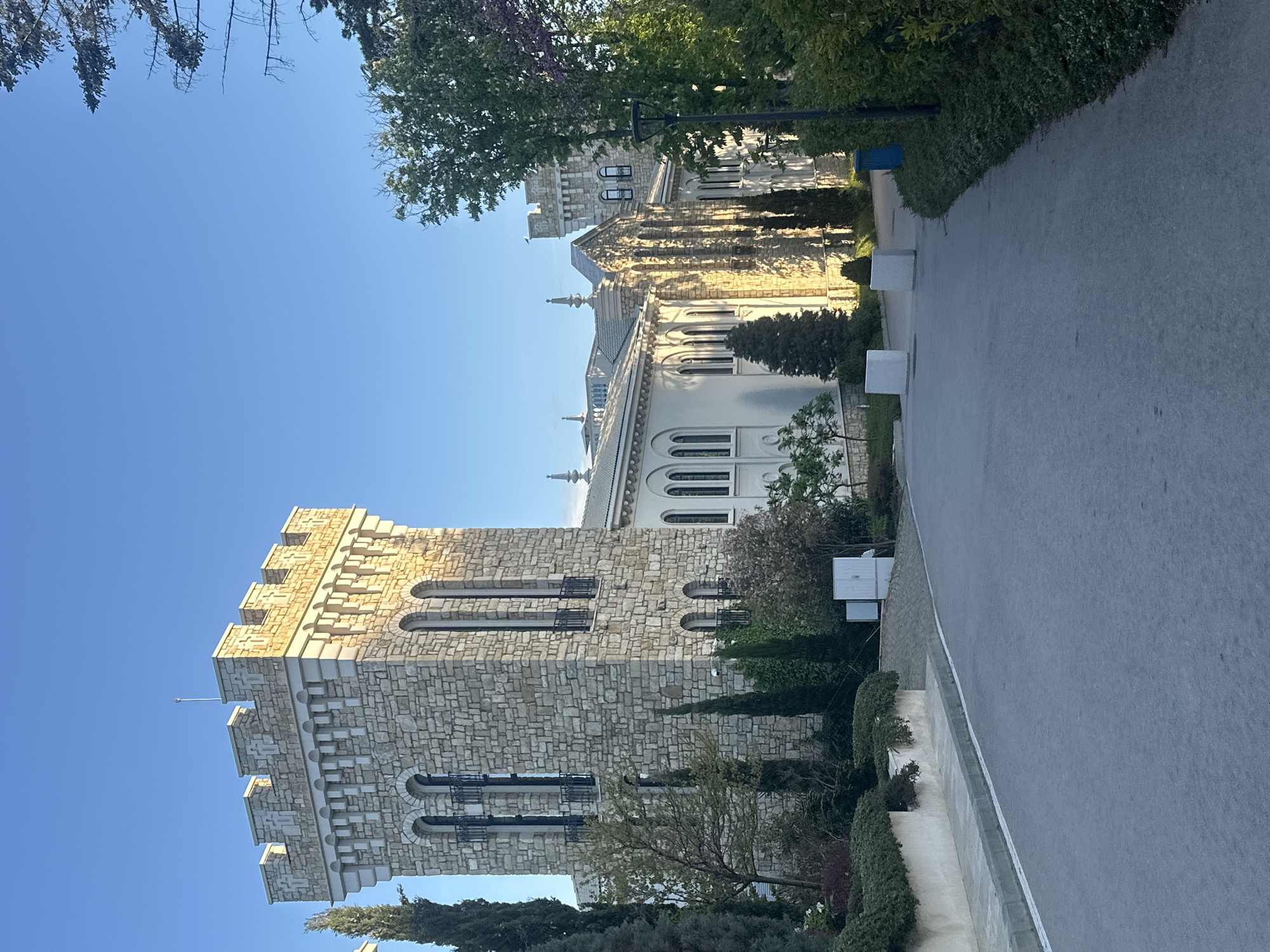 The image shows a tall stone building with a tower in Varna, Bulgaria. The building appears to be made of light-colored stone and has several arched windows. The tower is crenellated. The architecture of the main building is elaborate, painted a light color, with multiple arched windows and decorative details. A paved walkway runs alongside the building. The sky is blue and clear. There are trees and bushes around the building and alongside the walkway.