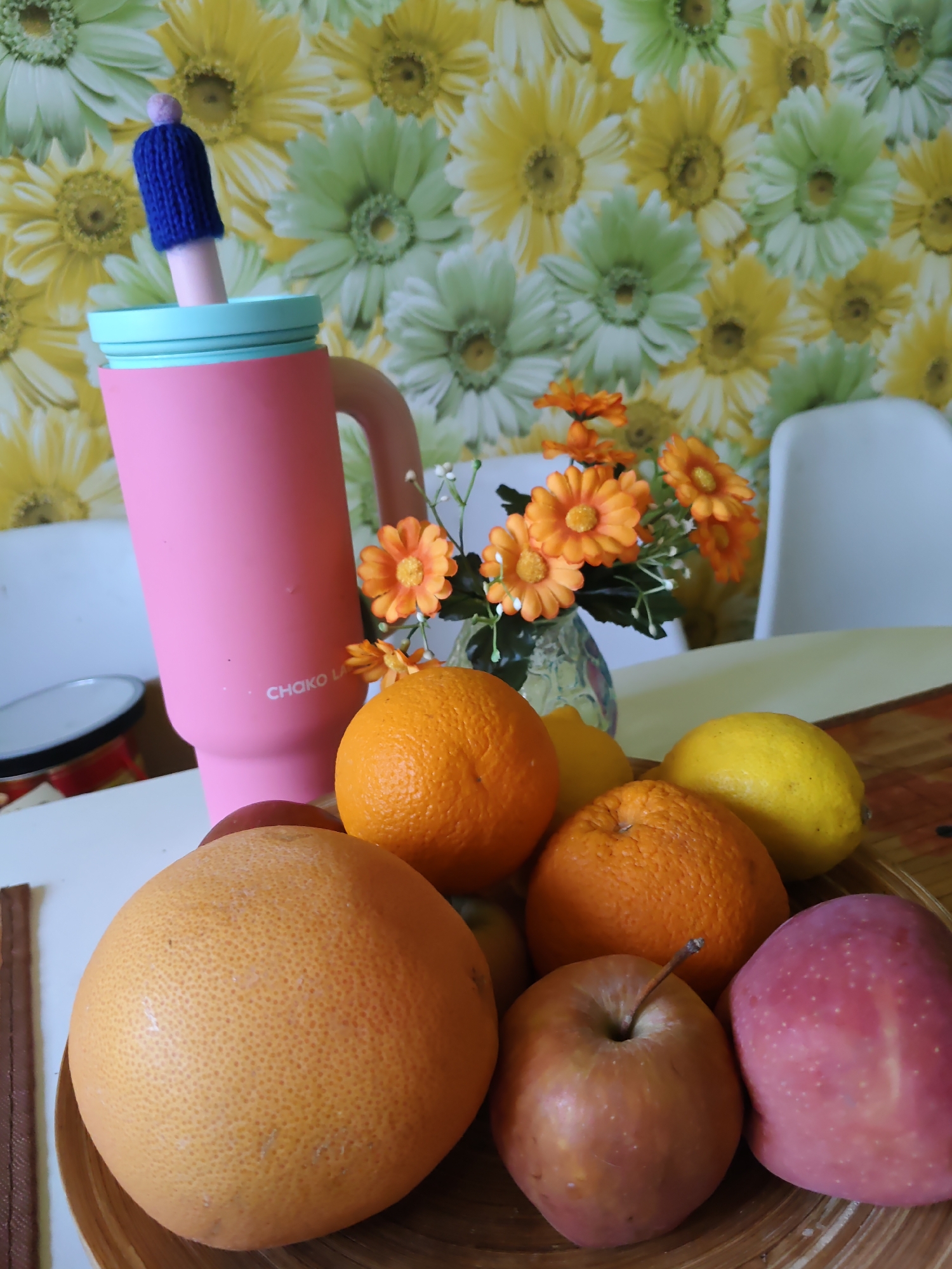 This is a still life photograph taken indoors, likely in a home environment, not on the streets of Rostov-on-Don, Russia. The image features a collection of fruits in a wooden bowl, positioned on a table with a patterned tablecloth. The fruits include a large grapefruit, two oranges, two lemons, two apples, and a few other smaller fruits. Behind the fruit bowl, a tall, pink insulated tumbler with a light blue lid and a handle is prominently displayed. A blue knitted cover with a small pom-pom adorns the straw protruding from the tumbler. In the background, a vase with artificial orange flowers and green foliage adds a decorative touch. The wall behind the vase is covered with floral wallpaper featuring a pattern of yellow and green daisies. Two white chairs are partially visible in the background, suggesting a dining or living area. The lighting appears to be natural, possibly from a nearby window, casting soft shadows on the scene. There are no people present in the image.