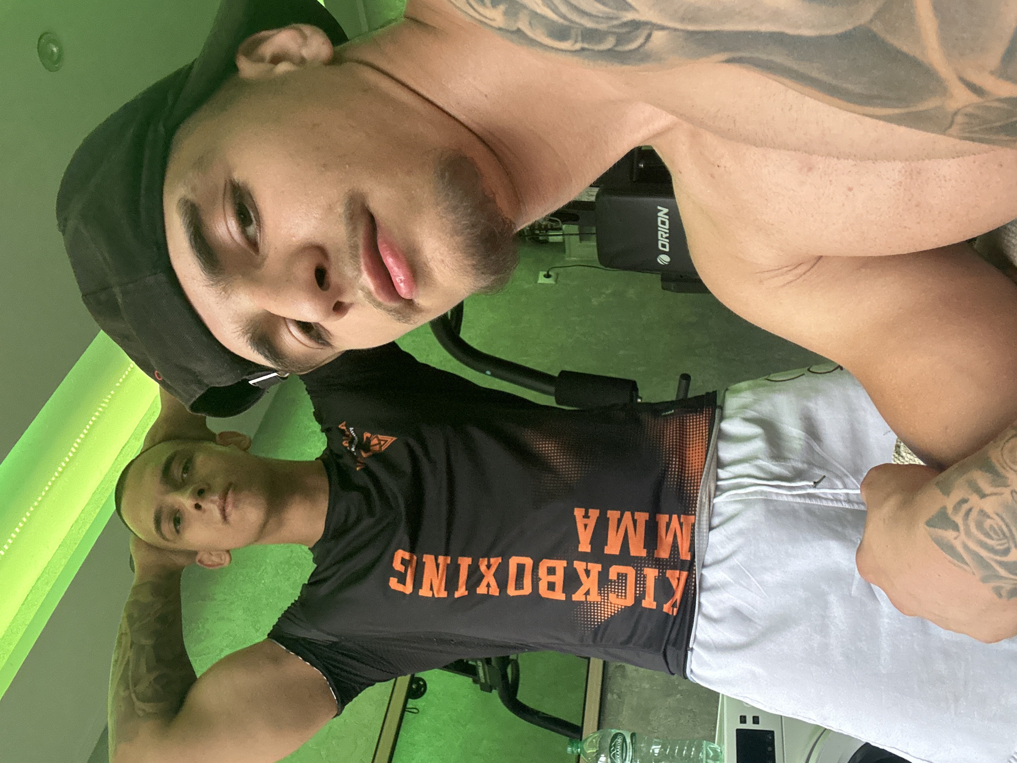 The image captures two males posing in what appears to be an indoor fitness setting, characterized by a distinct green illumination on the walls and floor. The photograph is rotated 90 degrees clockwise.
In the foreground, one male is featured in a close-up, looking directly at the camera with a neutral expression, showing a hint of a smirk. He has short dark hair, a dark baseball cap worn backward, and a short beard or heavy stubble. Visible tattoos adorn his arm and shoulder, suggesting a well-built physique. He appears to be shirtless or wearing a minimal garment that is not fully visible.
Behind him and to the left, the second male is seen from the waist up, also looking towards the camera with a neutral gaze. He has a very short haircut or buzz cut and visible tattoos on his arms. He is wearing a black t-shirt with prominent orange text that reads "KICKBOXING MMA" (appearing upside down due to the image's rotation). He is also wearing light-colored shorts.
In the background, a piece of exercise equipment with the brand name "Orion" is partially visible. A plastic water bottle can also be seen near the floor. The activity appears to be a casual pose for a photo. The environment suggests a gym or a dedicated workout space, highlighted by the artificial green lighting.