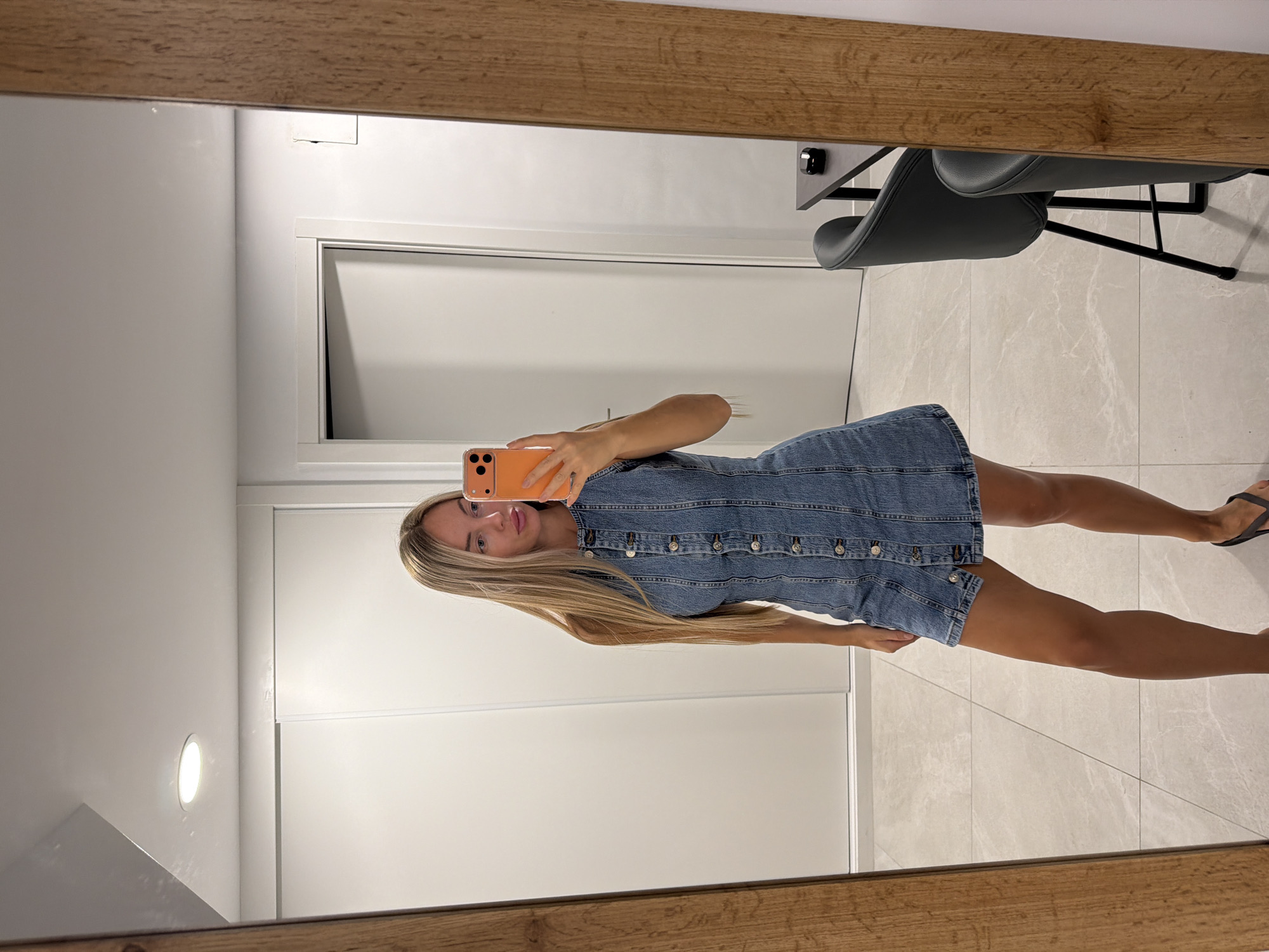 A woman is taking a mirror selfie in what appears to be a modern apartment. She is standing and wearing a denim dress. Her hair is long and blonde. She is holding an orange smartphone to take the picture. The background shows a tiled floor, a white wall with a circular light fixture, and a door. A wooden frame is visible in the foreground, which is part of the mirror. In the reflection, part of a desk and two chairs can be seen, along with a wall that is partially paneled. The scene suggests it is indoors, likely during the day. There is no visible text in the image. The overall impression is casual and everyday.