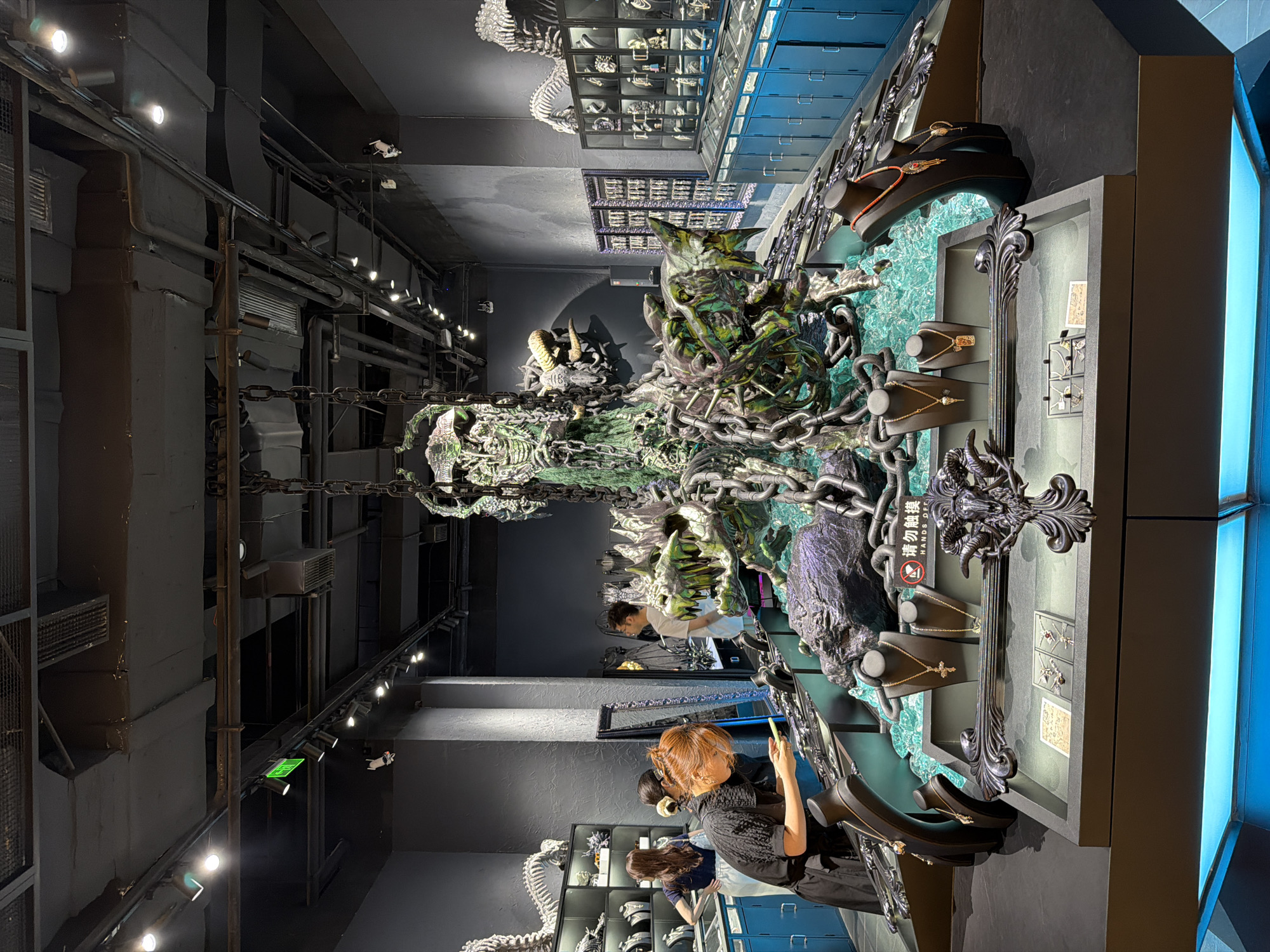 This image captures the interior of a unique jewelry store or exhibition space located in Yuzhong District, China. The central feature is a large, elaborate sculpture resembling a skeletal or decaying dragon, predominantly dark green and metallic, with substantial chains draped around and suspending parts of it from the industrial-style ceiling. The base of the sculpture is integrated into a multi-tiered display counter filled with fragmented clear-blue material, evoking water or ice.
In the immediate foreground, a woman with reddish-brown hair, wearing a black short-sleeved dress, leans over the display counter, engrossed in her mobile phone. To her left, partially obscured, another woman with dark hair in a blue top and white skirt examines merchandise in a glass display case against a dark textured wall. Further back on the right, a man with glasses and a white t-shirt stands in an aisle, observing displays.
The central display counter showcases various pieces of jewelry, primarily necklaces with ornate pendants (including crosses and skull motifs), presented on black busts and in flat display trays. A circular red "Please do not touch" sign is prominent near the sculpture. Along the left wall, shelving units feature additional jewelry and decorative items, topped by white skeletal models resembling dinosaur or dragon bones. The right wall is lined with multiple glass display cases exhibiting more jewelry, with rows of bright blue locker-like cabinets beneath them. The ceiling is exposed, revealing dark pipes, ducts, and numerous track lighting fixtures, along with a green exit sign. The floor is dark and unadorned. The overall atmosphere is dark, industrial, and distinctly gothic.