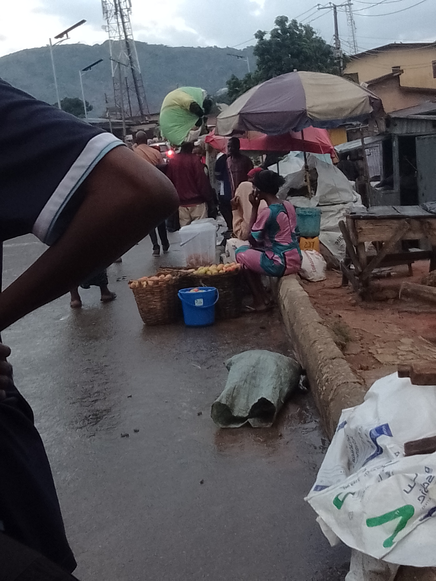 The image depicts a bustling marketplace scene in Gwong, Nigeria. In the foreground, a person's arm and shoulder are visible, suggesting they are either a participant in the market or a photographer. A young woman is seated on a log, dressed in a pink and blue patterned dress, appearing to be engaged in a transaction or conversation. In front of her are woven baskets filled with fruits, and a blue bucket. The ground is wet, indicating recent rain. Several other individuals are visible in the background, some carrying items on their heads, including one person with a green and yellow item on their head. The market is set up with stalls covered by umbrellas, and various goods are displayed. A telecommunications tower is visible in the distance, along with hills and buildings. The overall atmosphere is one of active commerce and daily life in a Nigerian urban setting.