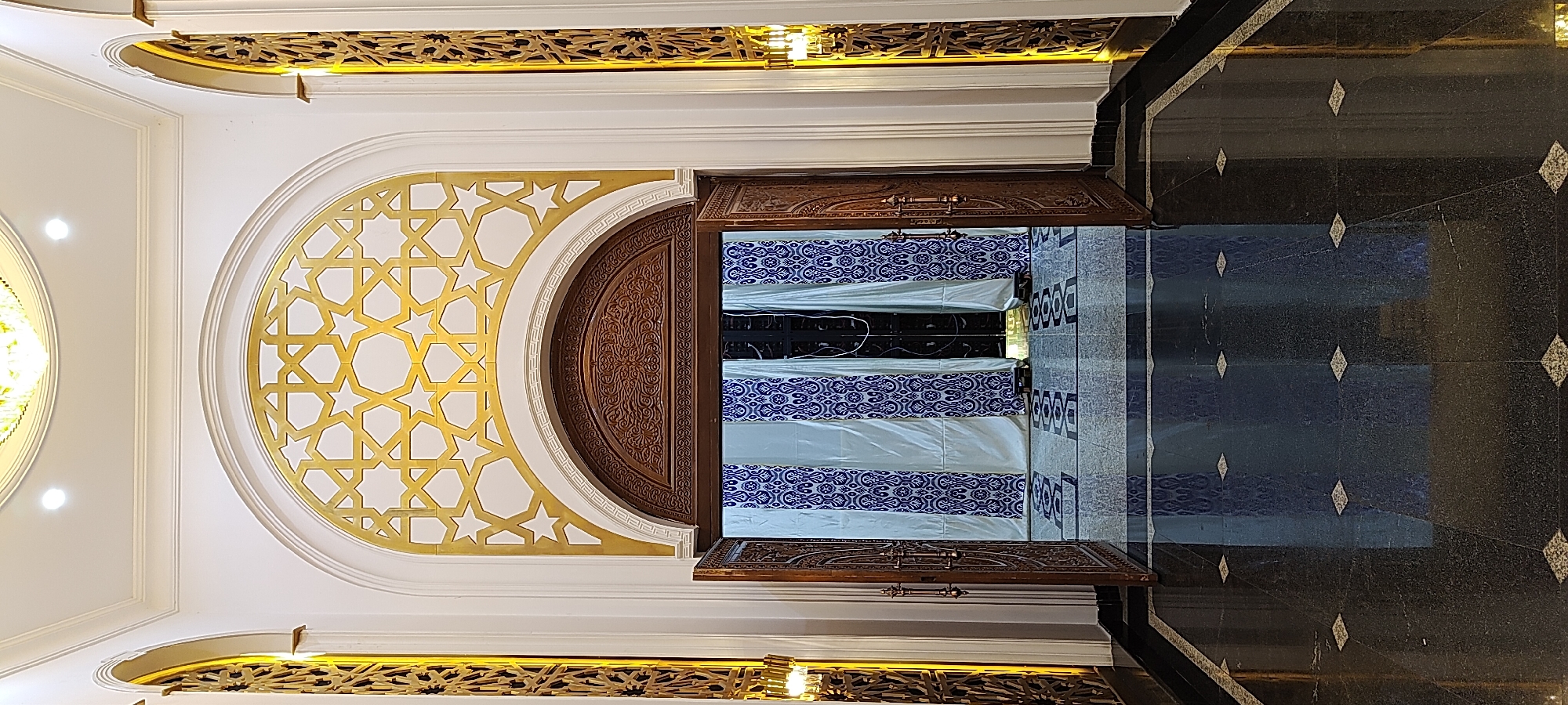 The image captures an ornate entryway, likely within a building in Khujand, Tajikistan, characterized by its Islamic architectural influences. The focal point is a large, arched wooden double door, partially open, revealing a backdrop of white curtains adorned with blue geometric patterns. Above the doorway, a golden geometric lattice design fills a segmental arch. The surrounding walls are white, and flanking the central arch are tall, narrow arched openings containing intricate gold-colored lattice work, illuminated from behind by warm light, suggesting decorative screens or windows. The floor is polished black granite with a reflective surface, inlaid with diamond-shaped lighter colored tiles. The overall impression is one of grandeur and sophisticated design. There are no people visible in the image. The scene is static and appears to be an interior architectural detail.