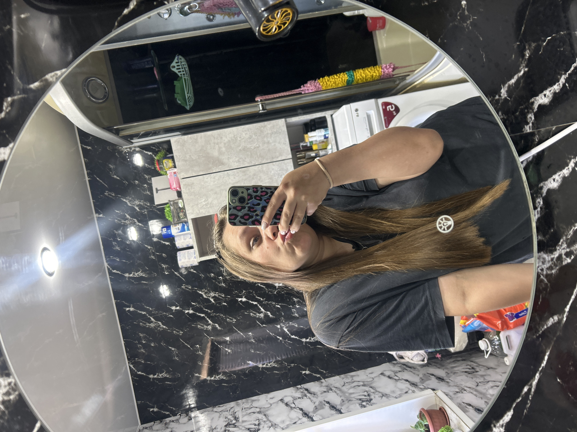 A person is taking a selfie in a circular mirror. The person has long brown hair and is wearing a dark t-shirt. They are holding a phone with a leopard print case. The background appears to be a bathroom with black marble walls, a white ceiling with a light fixture, shelves with various items, and a washing machine. A hairdryer is visible to the right. The location is specified as Olkhovka, Russia.