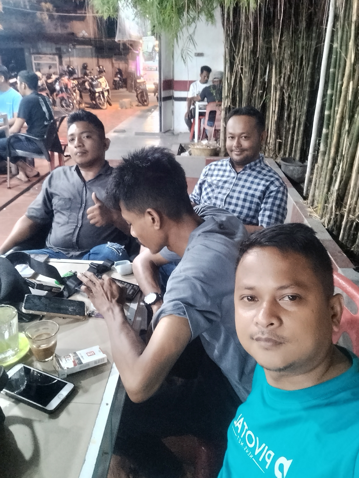 This media file captures a group of men socializing at what appears to be an outdoor or semi-outdoor cafe or eatery in Lhokseumawe, Indonesia, at night.
In the foreground, a man in a teal t-shirt takes a selfie, looking directly at the camera with a neutral expression. His t-shirt has white text that appears to be "PJVOTIA" (likely mirrored) and "MEMZ NET". Behind him, seated at a table, are three other men. To the left, a man in a dark grey shirt gives a thumbs-up and smiles. Next to him, a man in a light grey t-shirt is engrossed with his phone or objects on the table. Further back and to the right, another man in a blue and white checkered shirt smiles towards the camera. In the background, two more men are partially visible: one in a light blue t-shirt on the far left, and another in a white t-shirt further back on the right.
The table in front of the men holds several items: glasses with drinks (one green, one coffee-colored), multiple mobile phones (including a white iPhone and dark smartphones), a white pack of "Esse Change Double" cigarettes, and a watch. Behind the group, a wall of bamboo stalks creates a natural barrier or decoration. Beyond that, a street or parking area is visible with several motorbikes, some with riders, suggesting a bustling environment. The scene is illuminated by artificial lights, indicating it is nighttime. The overall atmosphere appears casual and relaxed, with the men engaging in social interaction.