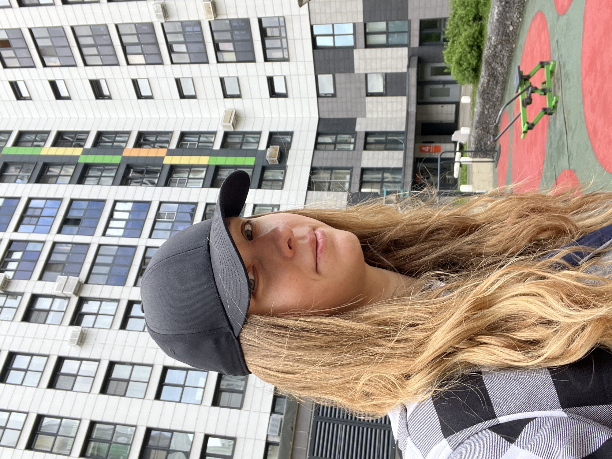 A vertically oriented, close-up shot depicts a young woman with long, blonde, wavy hair wearing a dark gray baseball cap and a black and white checkered shirt. She is looking towards the right side of the frame with a slight smile.
In the background is a multi-story building with numerous windows. The building's facade is a combination of white, gray, and blocks of color including blue, green, and yellow. Attached to the building are what appear to be air conditioning units.
Further in the background, slightly out of focus, is a playground with a green and red surface and play equipment. A strip of greenery separates the building and the playground area. The picture suggests an urban environment, likely in Mytishchi, Russia, although specific identifiable landmarks are not visible.