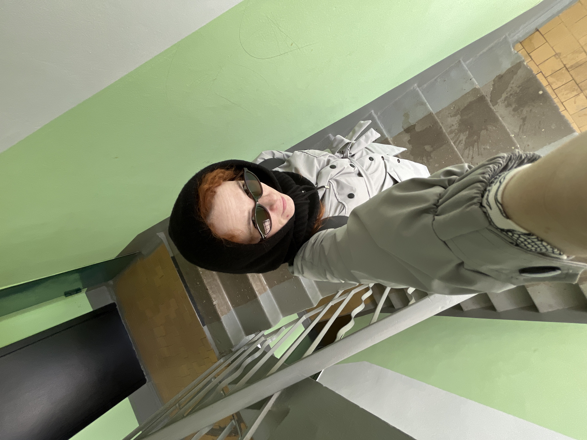 A low-angle, top-down selfie captures a person looking directly upwards at the camera from a staircase in an indoor setting, likely in Chaikovsky, Russia. The individual has reddish-brown hair, partly covered by a black hood or scarf, and wears dark sunglasses. They are dressed in a light grey, possibly quilted, jacket with visible black buttons down the front and a patterned cuff on the right sleeve.
The setting is a concrete staircase with worn, grey-painted steps that show signs of use and some dark splatters or stains. A grey metal handrail with multiple thin vertical bars runs along the side of the stairs. The walls feature a distinct two-tone paint scheme: the lower section is a vibrant light green, while the upper section is a lighter grey or white, divided by a sharp diagonal line. Minor cracks and scuff marks are visible on the wall surfaces. At the bottom of the stairs, a segment of yellowish-brown tiled flooring is visible alongside a dark, possibly black, door. The perspective emphasizes the descent of the staircase. No visible text is present in the image.