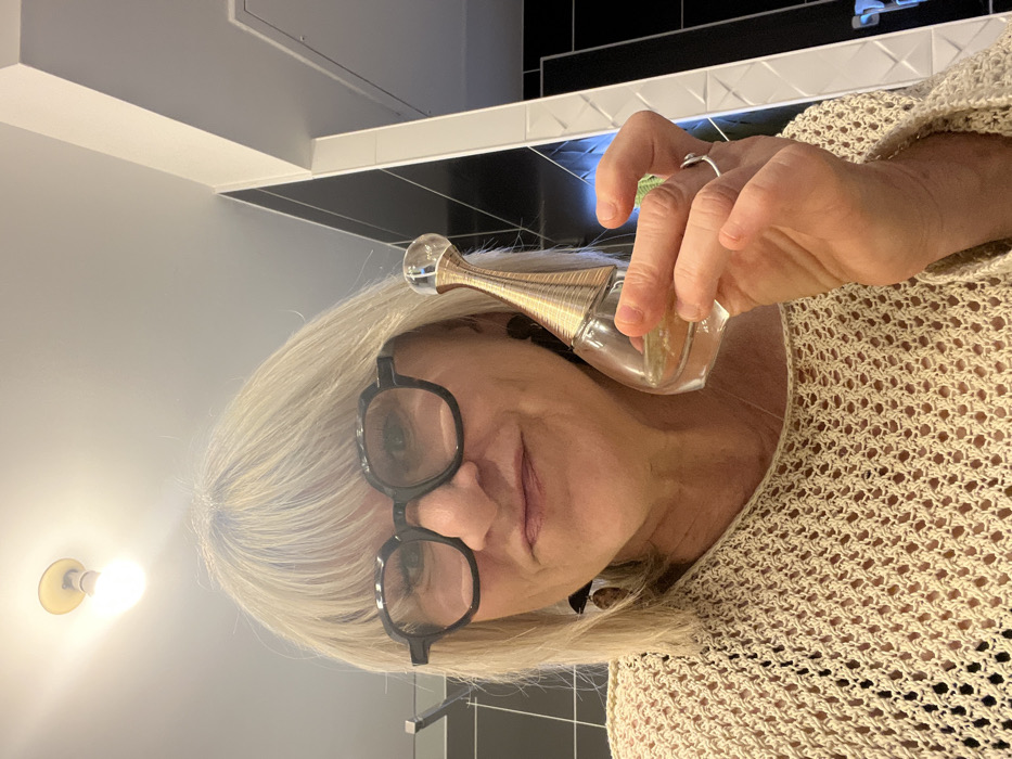A woman with blonde hair and black-framed glasses holds a Dior J'Adore perfume bottle up to her neck. She's wearing a beige knitted sweater. The background features a wall and tiled surfaces, with a light fixture visible on the left. Her left hand, with a silver ring, is visible.