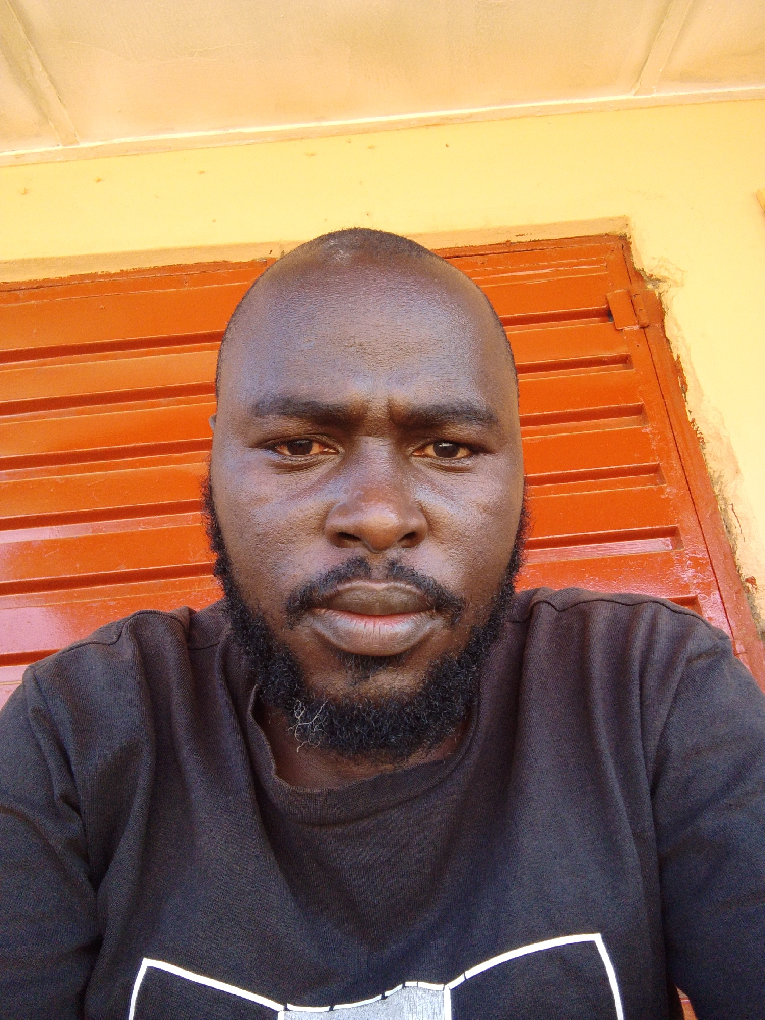A close-up photograph depicts a dark-skinned man with a shaved head and a full beard, positioned against a backdrop of orange horizontal panels and a light yellow wall. The man is wearing a dark-colored shirt. The location is possibly in the city of Jos, Nigeria.