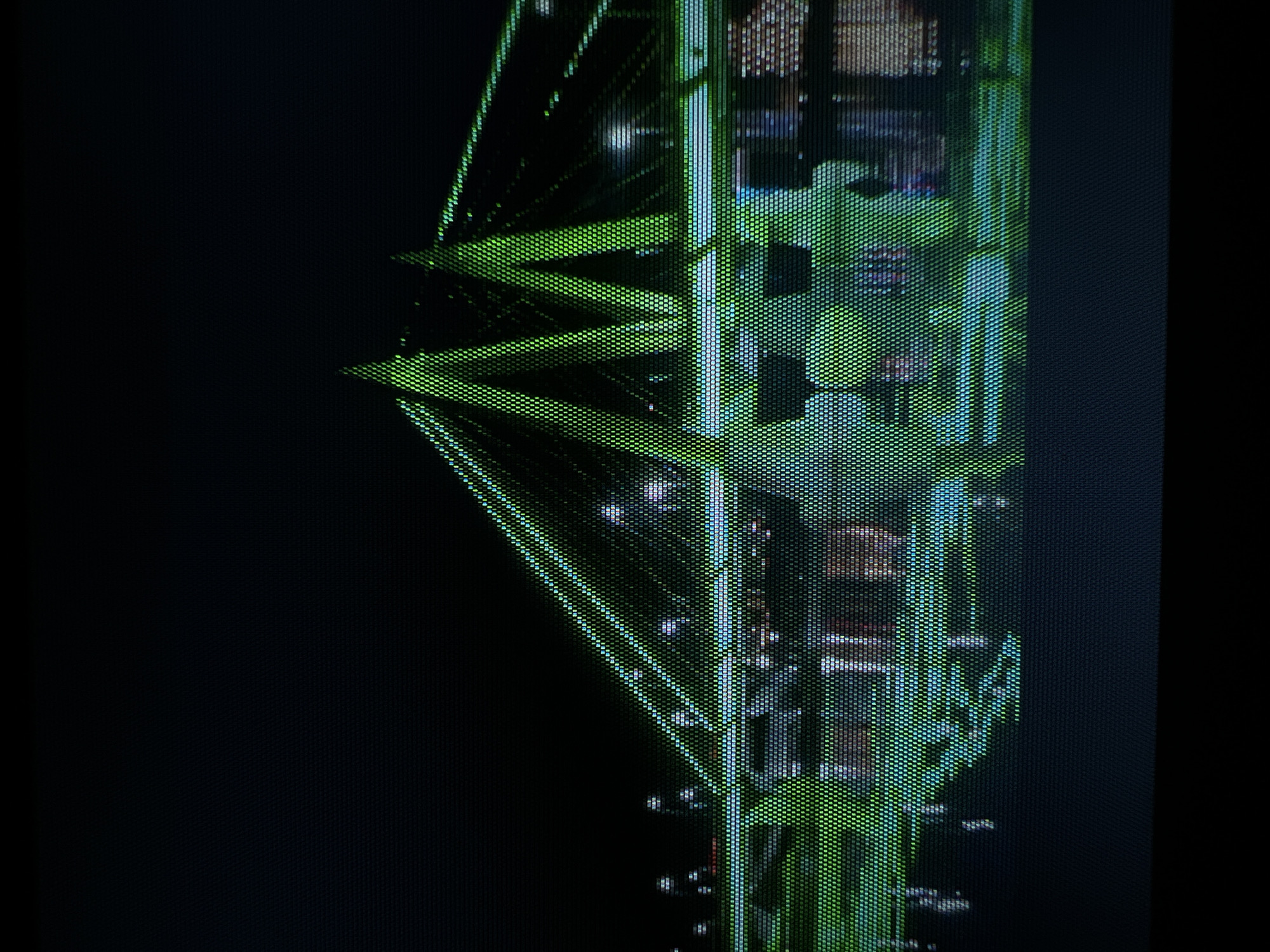 This media file captures a close-up, highly pixelated view of an illuminated architectural structure at night, dominated by bright green lights against a dark background. The most prominent feature is a large, distinctive "M" shape, formed by numerous individual light sources, suggesting an LED display or an extensively lit structural element.
Behind and around this central green "M," additional structural elements are visible, also composed of a grid-like pattern of lights in shades of green, blue, and white. These elements appear to be part of a larger construction, possibly bridge cables, supports, or intricate paneling. The strong pixelation throughout the image indicates either a very close shot of an LED screen or the individual LED lights on a large outdoor installation.
Given the location context of Kazan, Russia, the prominent "M" shape strongly identifies the subject as the iconic **Millennium Bridge**, known for its distinctive M-shaped pylons and extensive night-time illumination. The image captures the bridge's lighting system up close, creating a textured, almost digital appearance. The dark background confirms it is night. There are no people, explicit activities, or visible text beyond the structural "M" symbol. The scene conveys a sense of modern, illuminated architecture.