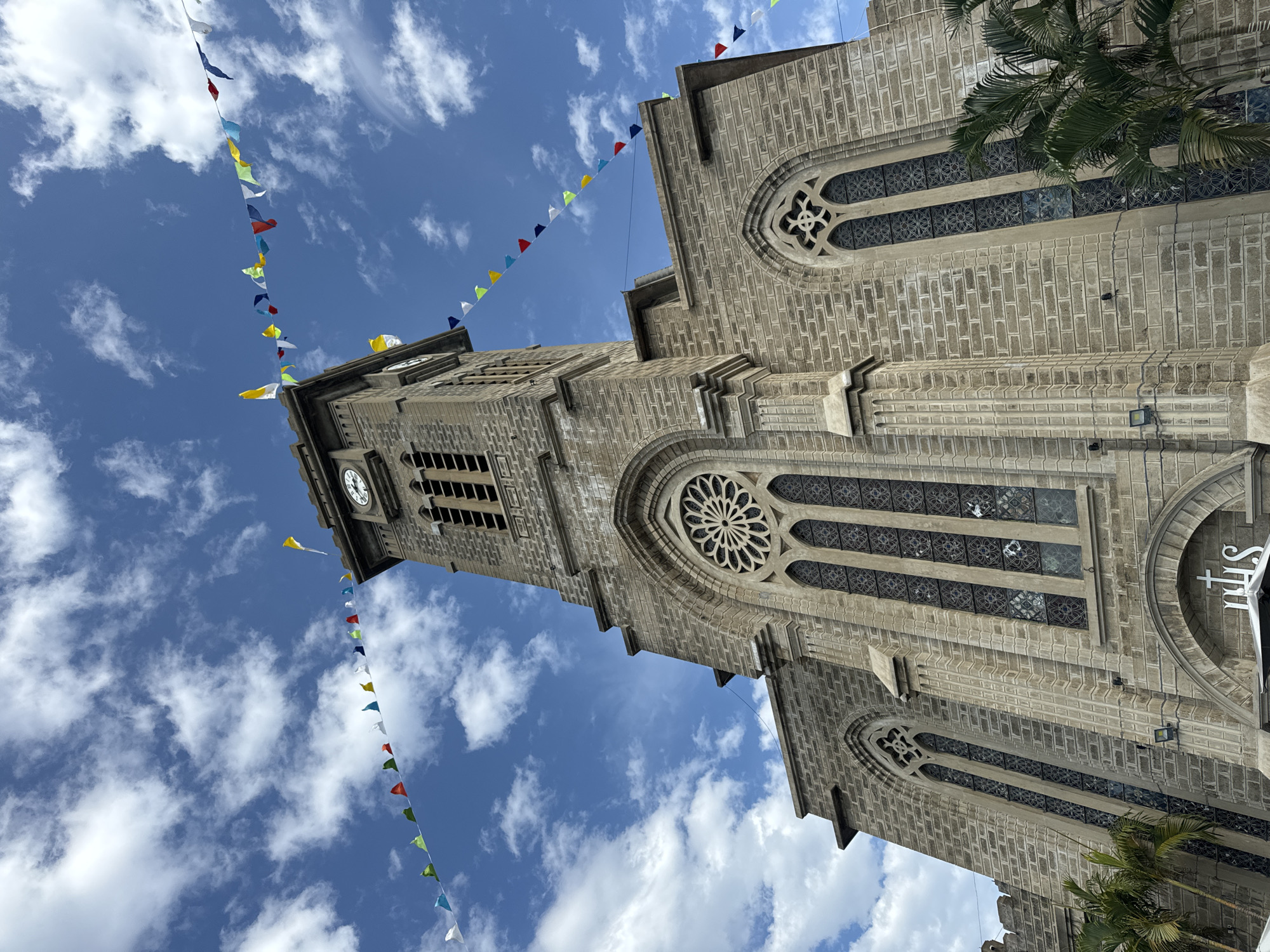 The image captures the front facade of the Nha Trang Cathedral in Nha Trang, Vietnam, under a bright blue sky dotted with white, fluffy clouds. The cathedral is constructed of grey stone bricks and features gothic architectural elements, including pointed arches, stained-glass windows with intricate patterns, and a prominent clock tower. A rose window is visible above the main entrance area. Colorful triangular flags are strung across the sky, adding a festive atmosphere to the scene. Palm trees are visible on the right side of the image, partially obscuring the lower part of the cathedral's facade. The camera angle is low, looking up at the cathedral, emphasizing its height and grandeur. There are no people or discernible activity in the image.