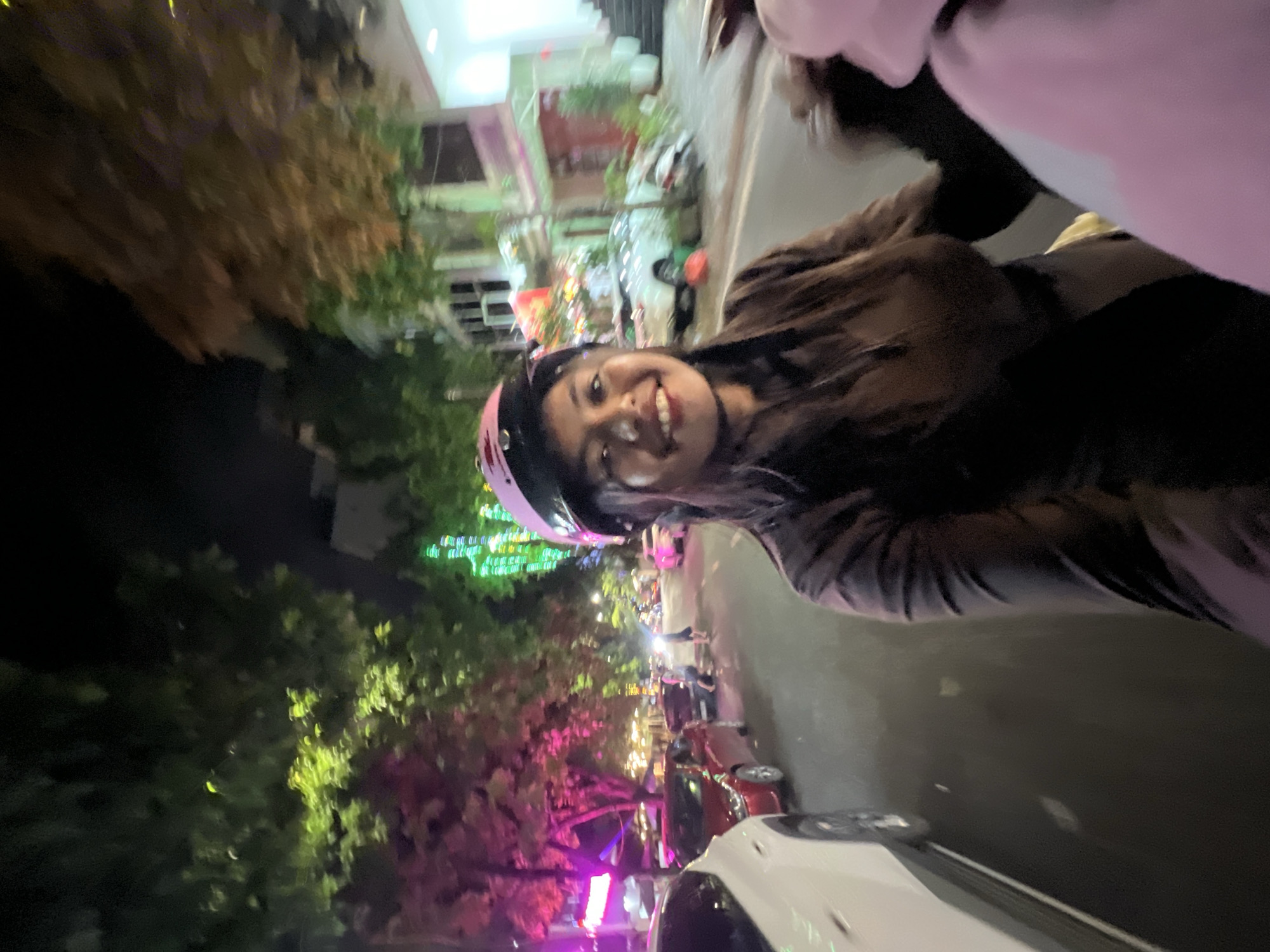 In a nighttime, low-light street scene in Minh Xuân Ward, Vietnam, a young woman with dark hair and a pink helmet smiles at the camera. She is wearing a dark brown jacket or shirt and is on a motorcycle. The background shows a street with cars, trees illuminated by pink and green lights, and buildings. The perspective suggests the photo was taken from another motorcycle or vehicle moving alongside hers.