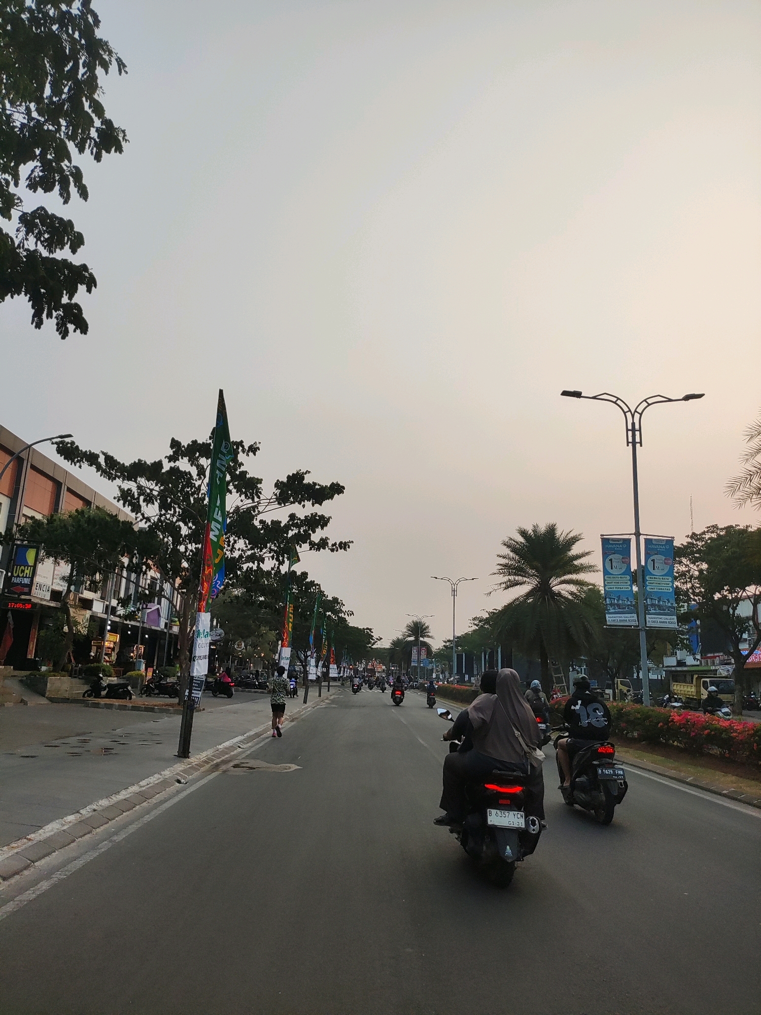 The image depicts a street scene in Telagaasih, Indonesia, likely during the late afternoon or early evening given the soft, diffused lighting and the gradient of the sky. The perspective is from a vehicle on the road, looking forward.
On the left side of the road, there is a row of commercial buildings with storefronts. Signs are visible, some in Indonesian, indicating businesses like "UCHI PARFUMERIE" and others. Trees line the sidewalk in front of these buildings, and several flags are mounted on poles along the street.
The road itself is paved and appears to be a multi-lane thoroughfare. Several motorcycles are in motion, with riders. In the foreground, two motorcycles are prominent. The one on the left has a rider and a passenger, both wearing dark clothing, and the license plate reads "B 6357 YCN". The motorcycle to its right has a single rider wearing a black hoodie, with the license plate "B 5629 FRB". Further down the road, more motorcycles and a few cars can be seen in the distance.
To the right of the road, there is a sidewalk and then a landscaped area with trees, including palm trees, and flowering bushes. Streetlights are visible, including a prominent one with a double lamp fixture. Two large banners are displayed on poles near the streetlight, featuring promotional material with the text "HAVANA".
A lone individual is seen jogging on the right sidewalk. The overall atmosphere suggests a typical urban street with active transportation and commercial activity. The sky is a pale orange and white, indicating a possibly hazy or overcast condition, or perhaps the approaching sunset.
