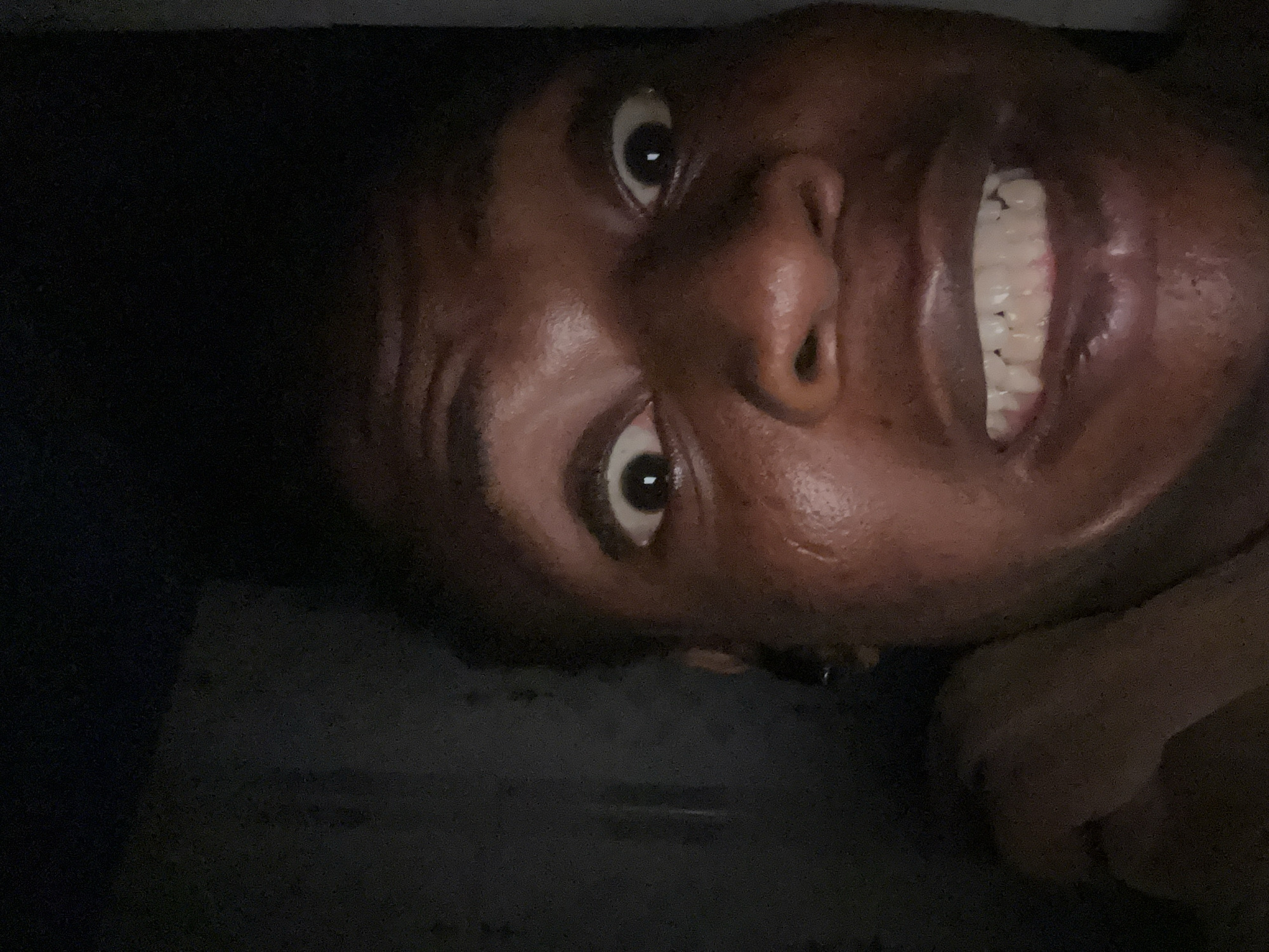 A close-up, slightly tilted image of a person's face with dark skin. The person is smiling broadly, exposing their teeth. The background is dark and indistinct. The location is Unknown, Unknown.