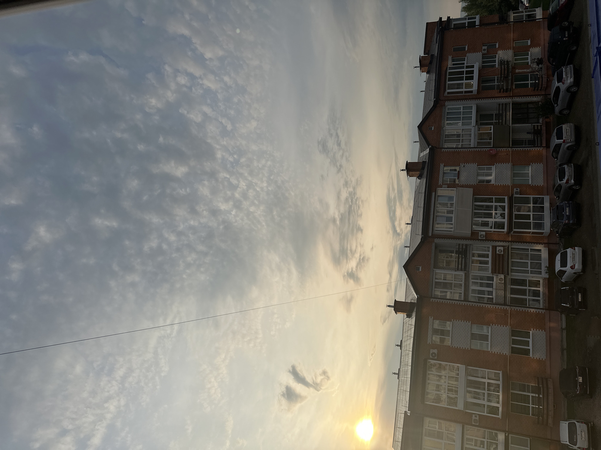 The image is a daytime, low-angle shot of a brick apartment building in Ussurijsk, Russia, under a cloudy sky. The building is multi-story and features numerous windows and balconies. Several cars are parked in front of the building. A thin wire runs diagonally across the sky. The sun is visible on the left side of the frame, casting a warm glow. The overall scene depicts an urban residential area.