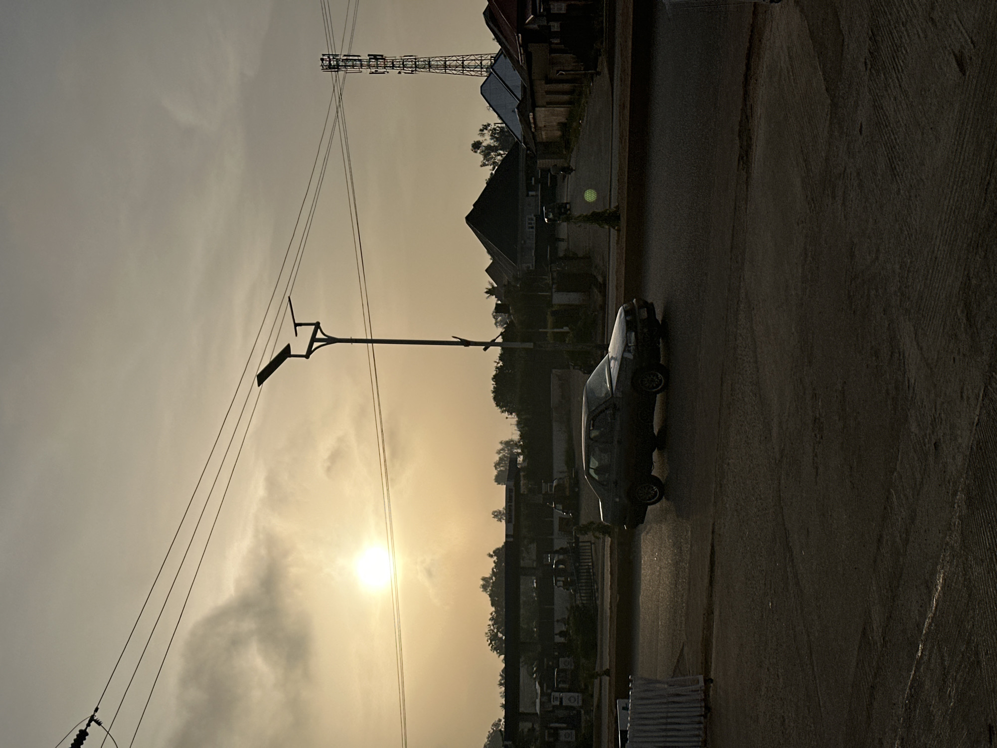 The image depicts a street scene in Bukuru, Nigeria at sunset. The sun is low in the sky, obscured by light clouds and haze. A black sedan is parked on the right side of the road. A utility pole with two streetlights attached is visible on the left side of the road. Power lines extend from the pole towards the right side of the image. In the background, buildings are visible, along with a cell phone tower. The street is paved and appears damp.