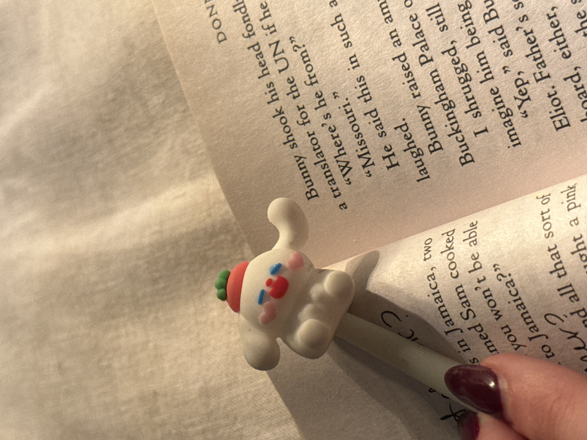 A close-up shot focuses on an open book with a cartoon rabbit pen resting on the pages. The text on the left page mentions "Jamaica" and "Sam," while the right page contains dialogue like "Bunny shook his head fondly," "translator for the UN," and "Missouri." Part of a hand with a maroon-colored nail is visible in the bottom right corner, holding the book. The lighting is soft, casting shadows and highlighting the book's texture. The setting is undefined, with only a glimpse of off-white fabric in the background.
