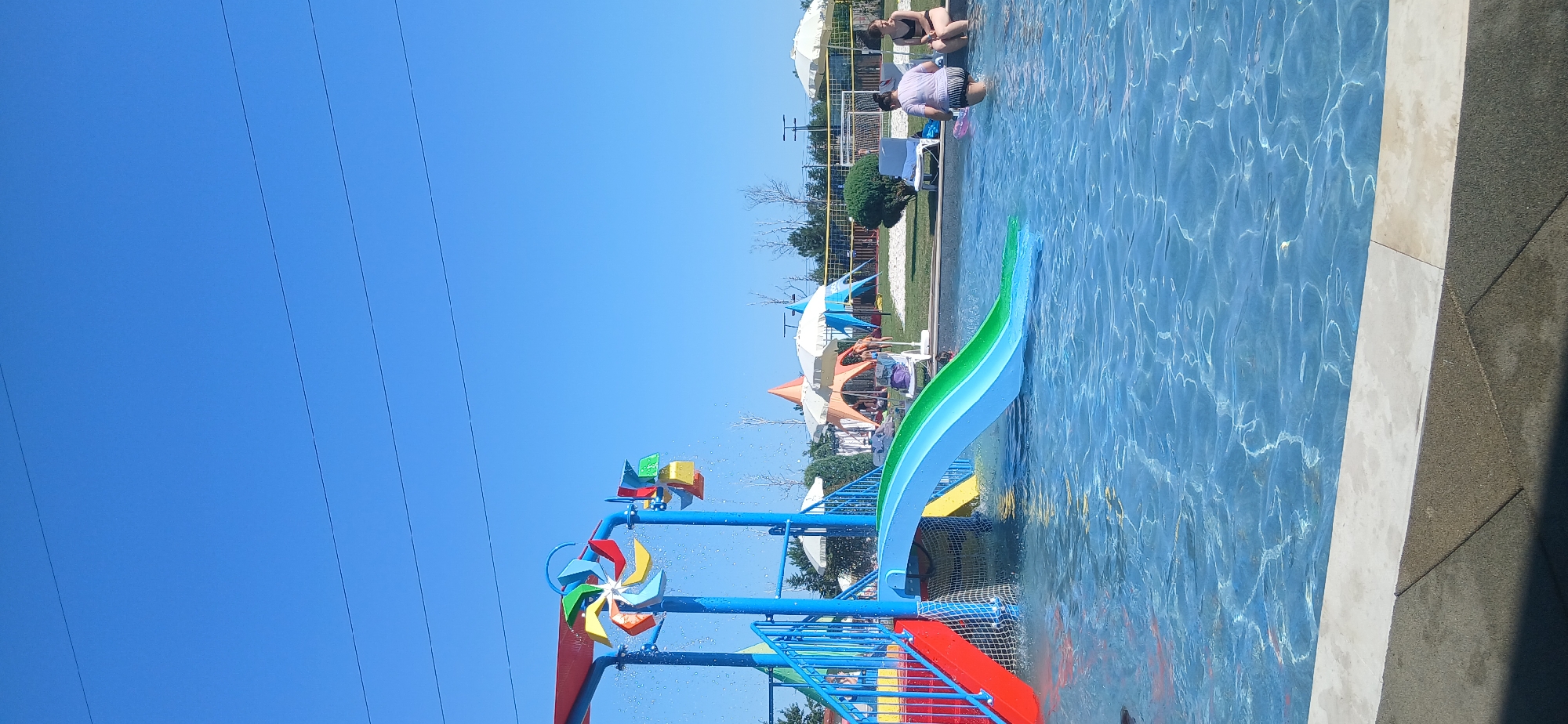 In an outdoor swimming pool area under a clear blue sky, a bright blue and red water slide complex is the central focus. The slide's curved, green and blue flume empties into the clear blue water. Colorful pinwheels with red, yellow, green, and blue blades are attached to the blue metal structure of the slide. In the background, several white and orange umbrellas provide shade over sand and grass areas. A volleyball net is visible in the distance. Two individuals are present near the edge of the pool: one person wearing a purple t-shirt and striped shorts is standing in the water, while another person wearing a black bikini is sitting on the pool's edge. The water reflects the sunlight, creating shimmering patterns on its surface.