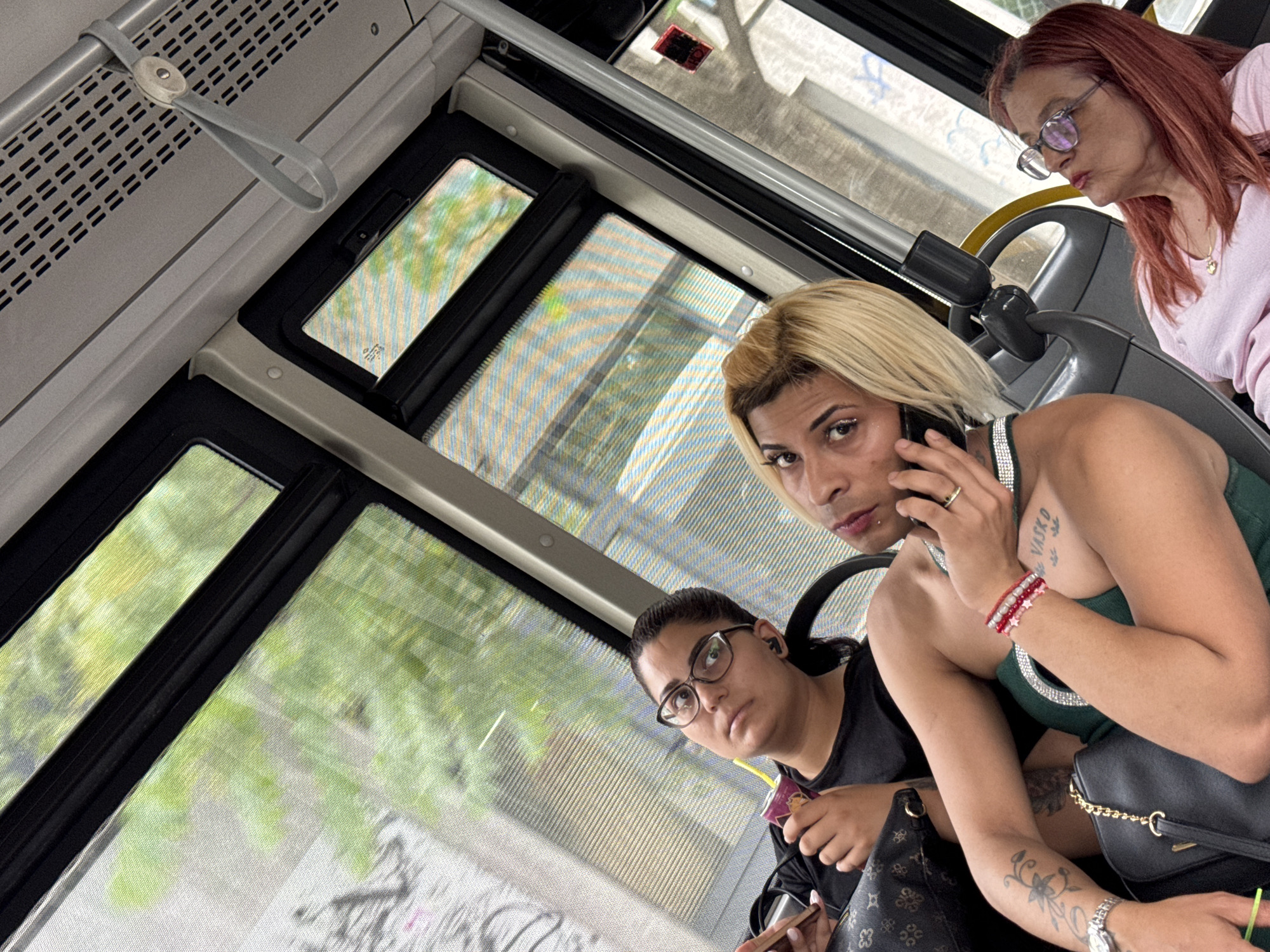 The image shows three people inside a bus. The person in the foreground, with blonde hair and makeup, is looking at the camera while holding a phone to their ear. They are wearing a green top with decorative straps and have tattoos on their arm and chest, with the word "VASKO" visible on their chest. They are also wearing bracelets on their wrist. To the left, a person with dark hair and glasses is looking upwards and to the left. They are wearing a black shirt and have earbuds in. They are holding a small box with a straw. To the right, a woman with reddish-brown hair and glasses is looking downwards. She is wearing a light pink top and a necklace. The bus has windows showing a blurry green outdoor scene, suggesting it is daytime and possibly a sunny day with foliage visible. The interior of the bus includes handrails and seats. The overall mood appears to be casual, with the individuals engaged in their own activities or interactions. The location is identified as Sofia, Bulgaria.