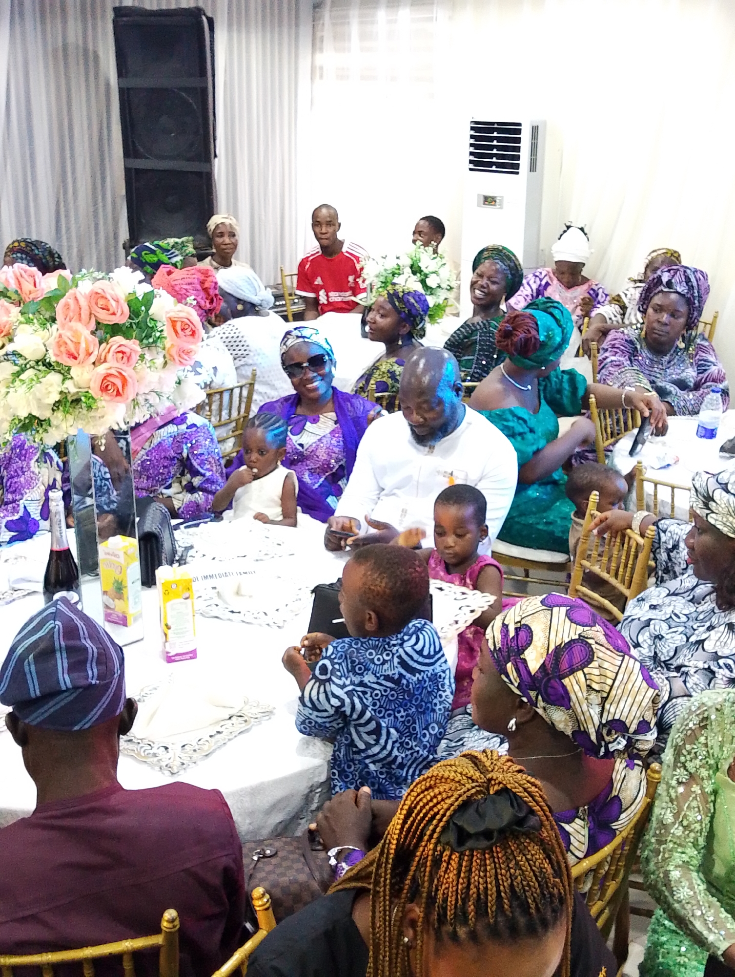The image captures a gathering of people seated around tables indoors. This is likely a special event. Many of the people are wearing traditional Nigerian attire. In the foreground, there are people seated at a table covered with a white tablecloth. On the table, there are items such as juice boxes, a bottle, and other objects. A large floral arrangement is present on one of the tables. The people are interacting with each other. A large speaker is visible in the background. Based on the location information provided, this scene is likely taking place in Kubwa, Nigeria.