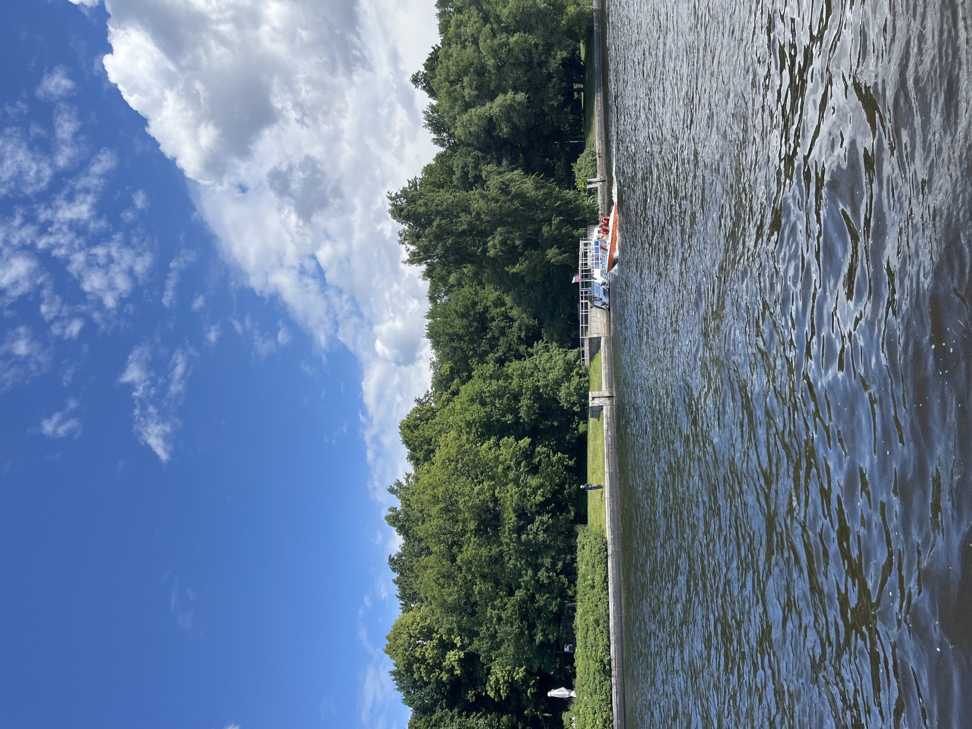 The image captures a view of the Neva River in Saint Petersburg, Russia, under a partly cloudy blue sky. In the foreground, dark blue water with visible ripples occupies the lower half of the frame. A narrow strip of a stone embankment runs along the middle ground, backed by a lush green lawn and dense trees that form a thick wall of foliage.
On the embankment, a person in dark clothing stands near the water's edge, facing away from the viewer. Further to the right, a small, orange motorboat with several occupants wearing life vests speeds across the water, leaving a white wake behind it. Adjacent to the motorboat, a larger white boat is docked at a white pier. A small Russian flag is visible on the pier.
The shoreline extends to the left, featuring a large white umbrella, a white gazebo-like structure, and more manicured greenery. The overall impression is a serene natural setting within an urban environment, characterized by the contrast between the calm water, verdant trees, and the presence of recreational boats.