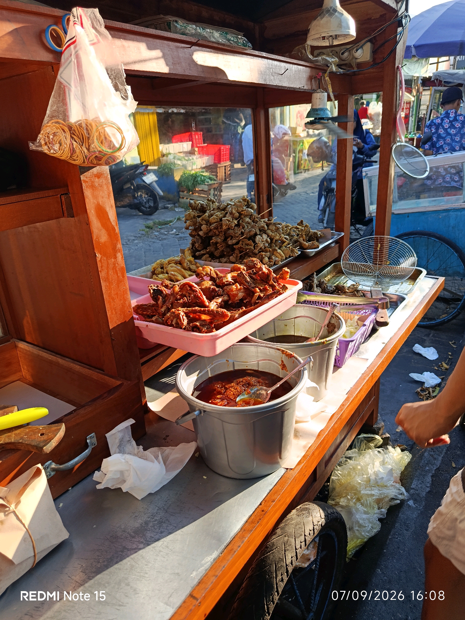 This is a street-level photograph taken in Keboansikep, Indonesia, on September 7, 2026, at 16:08. The image depicts a food stall with various fried food items, likely for sale. A wooden cart serves as the stall's structure. In the foreground, a portion of a person's right arm and leg are visible, suggesting they are a customer or vendor. The stall is laden with food. Two plastic buckets are prominent: one contains a dark, savory sauce, while the other holds a bright red chili sauce with a spoon. Above these, a pink tray is filled with pieces of fried chicken. Further back, skewers of what appear to be fried snacks are displayed on a metal tray. A metal sieve rests on another tray. A clear plastic bag filled with rubber bands hangs on the left side of the stall. The background shows a bustling street scene with other stalls, a motorcycle, and various individuals, some of whom are vendors. The lighting suggests it is late afternoon, with shadows cast across the scene. The overall impression is of an active street food vendor in a busy urban environment.