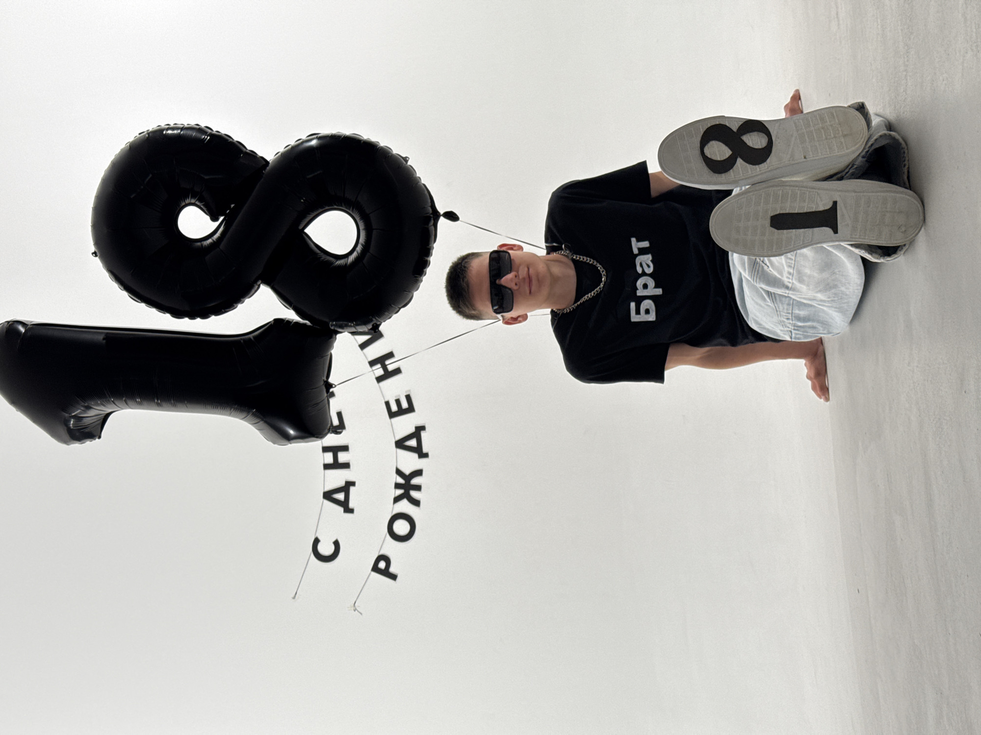 A young man is posed for a photo with balloons and a banner. He is wearing a black t-shirt with white Cyrillic text that reads "Брат" (Brother), sunglasses, and light-colored pants. His shoes have the numbers "1" and "8" on their soles, indicating his age or a significant number. Two large black balloons are shaped like the numbers "1" and "8," and a banner with the Cyrillic words "С ДНЕМ РОЖДЕНИЯ" (Happy Birthday) hangs between them. The background is a plain white studio setting. The lighting is bright, suggesting an indoor photoshoot. The overall mood appears celebratory and youthful. The location context is Chaikovsky, Russia.