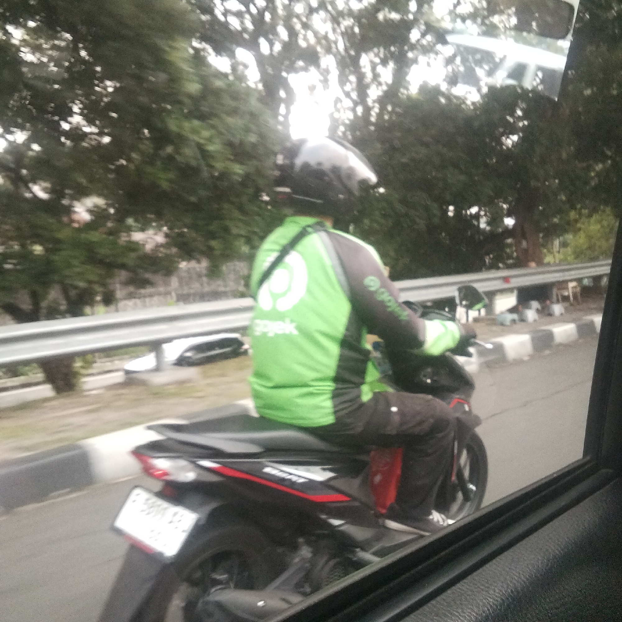 A person wearing a green and black Gojek jacket and helmet rides a black motorcycle with red accents on a paved road. The motorcycle has a white license plate. The rider is positioned in the right lane of traffic, moving away from the viewer. The scene appears to be in an urban or suburban environment with trees and foliage in the background, and a roadside barrier. The image is taken from inside a vehicle, as evidenced by the car window frame visible on the right side. The specific location within Bogor, Indonesia, cannot be determined from the provided image alone.