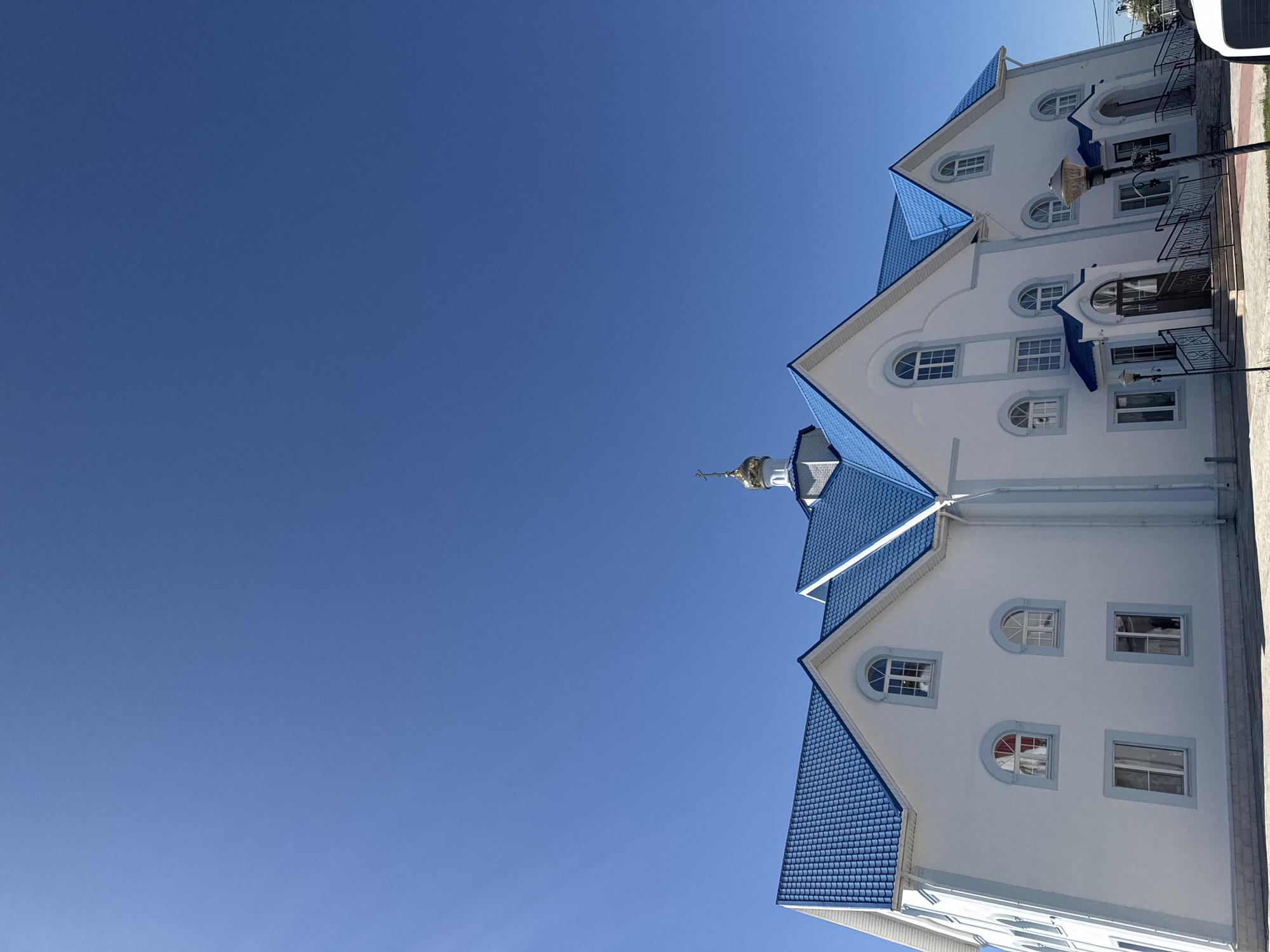 A low-angle, rotated view shows the side of a white, multi-story building with a steep, blue-tiled roof against a clear blue sky. The building is constructed with multiple gable-roofed sections. The windows are arched, with varying designs visible. A golden dome with a cross sits at the top of one section of the building. The building appears to be in the city of Unknown, Unknown.