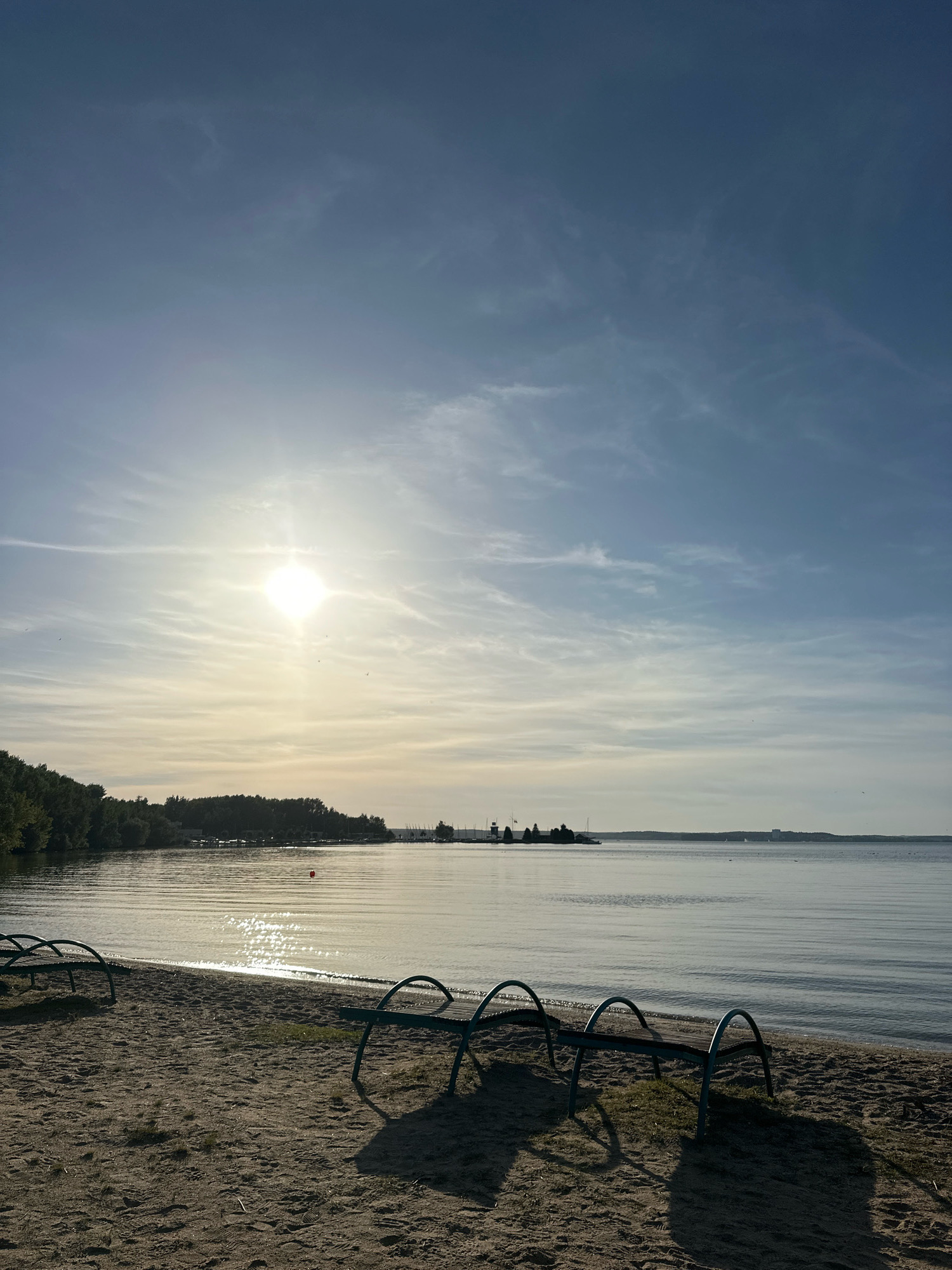 The image depicts a serene beach scene on a sunny day. The foreground is a sandy beach with a few empty, blue-accented benches casting long shadows. In the midground, the calm water of a lake or sea stretches towards the horizon, reflecting the bright sun. The sun is positioned high in the sky, slightly to the left of center, with wispy clouds scattered across the blue expanse. The background features a tree-lined shore with some buildings and structures visible in the distance, suggesting a populated area. The overall impression is one of tranquility and natural beauty, typical of a recreational area. The location is identified as Zhdanovichsky Selsoviet, Belarus.