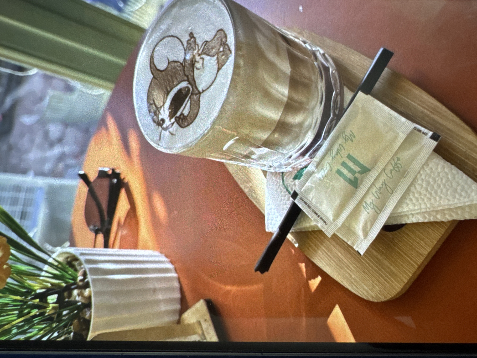 This image captures an indoor table setting, likely within a cafe, in Vratsa, Bulgaria. The central subject is a clear glass containing a layered coffee beverage, likely a latte, topped with detailed latte art. The art depicts a cartoon mouse character, specifically Jerry from "Tom and Jerry," with its mouth wide open in a screaming or yawning expression, rendered in shades of brown and white foam.
The glass rests on a light-colored, rectangular wooden tray with rounded edges. Alongside the glass, a black plastic straw with a visibly broken or jagged tip is present. Beneath the straw and partially under the glass are several folded white paper napkins with a textured, dimpled surface. Stacked on these napkins are two small, rectangular sachets, light green/beige in color, each bearing the text "My Way Caffe" and a stylized "M" logo.
The table surface is a glossy, reddish-orange color, reflecting ambient light. In the upper left background, a white fluted ceramic pot contains a green leafy plant with narrow, upright stalks and light-colored decorative stones or pebbles at its base. To the right of the plant pot and further into the background, a pair of dark-lensed sunglasses with black frames lies on the table, casting a distinct shadow. The scene is illuminated by warm, possibly natural light, creating reflections on the table and glass.