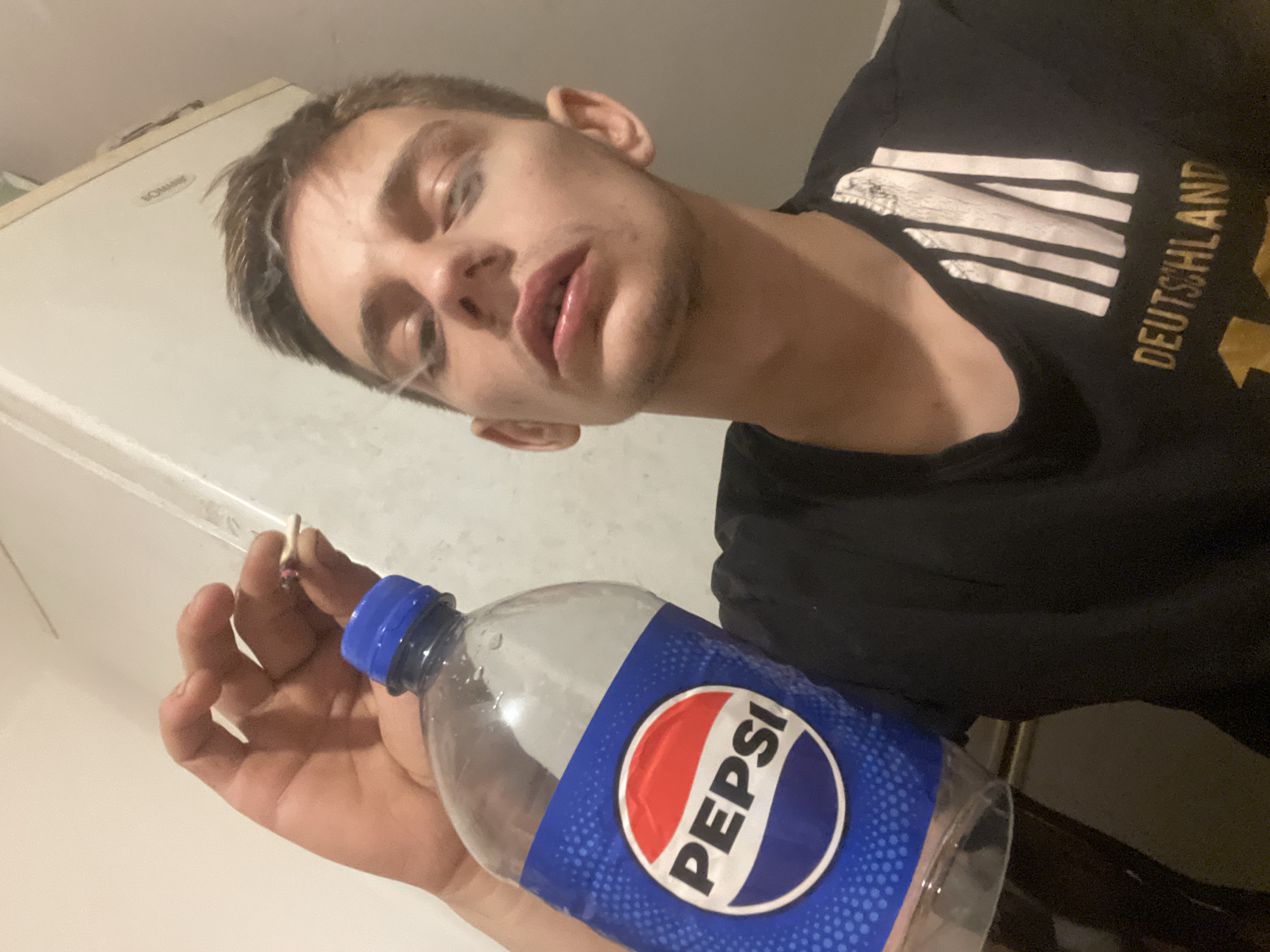A young adult is holding a bottle of Pepsi and smoking. The person is wearing a black t-shirt with white stripes on the sleeve and the word "DEUTSCHLAND" written in gold letters. The background appears to be an indoor setting with a white wall and a white appliance. There is a stream of smoke rising from the cigarette.