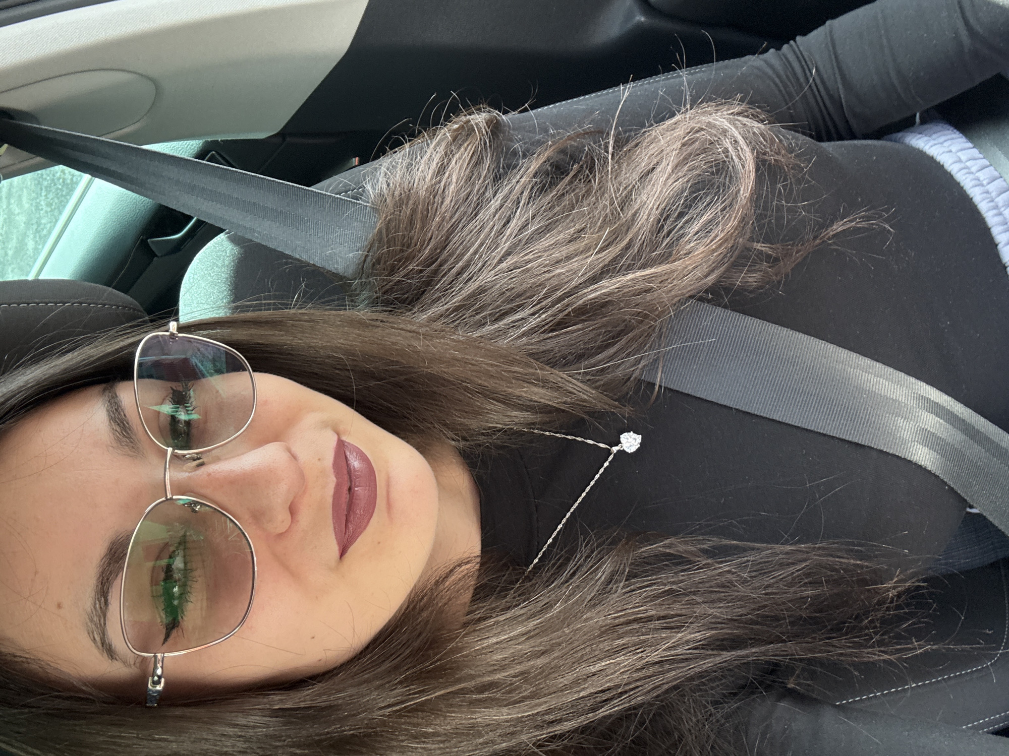 The scene depicts a close-up, frontal view of an adult female seated in the passenger compartment of a vehicle, located in Bad Buchau, Germany. The female, occupying the majority of the frame, has long, dark brown hair and is wearing eyeglasses with thin, metallic, possibly gold-toned frames and tinted lenses. Her lips are coated with a dark red or berry-toned lipstick. She is dressed in a black long-sleeved top and accessorized with a silver-toned chain necklace from which a clear, heart-shaped pendant is suspended. A black vehicular seatbelt is securely fastened across her chest, extending diagonally from her upper left shoulder to her lower right torso. Visible elements of the vehicle's interior include the top portion of a grey fabric headrest behind her and a light-colored door panel with black trim on the right side of the frame. A blurred, greenish-blue area, indicative of an exterior environment or reflection, is visible through the window in the upper right background.
