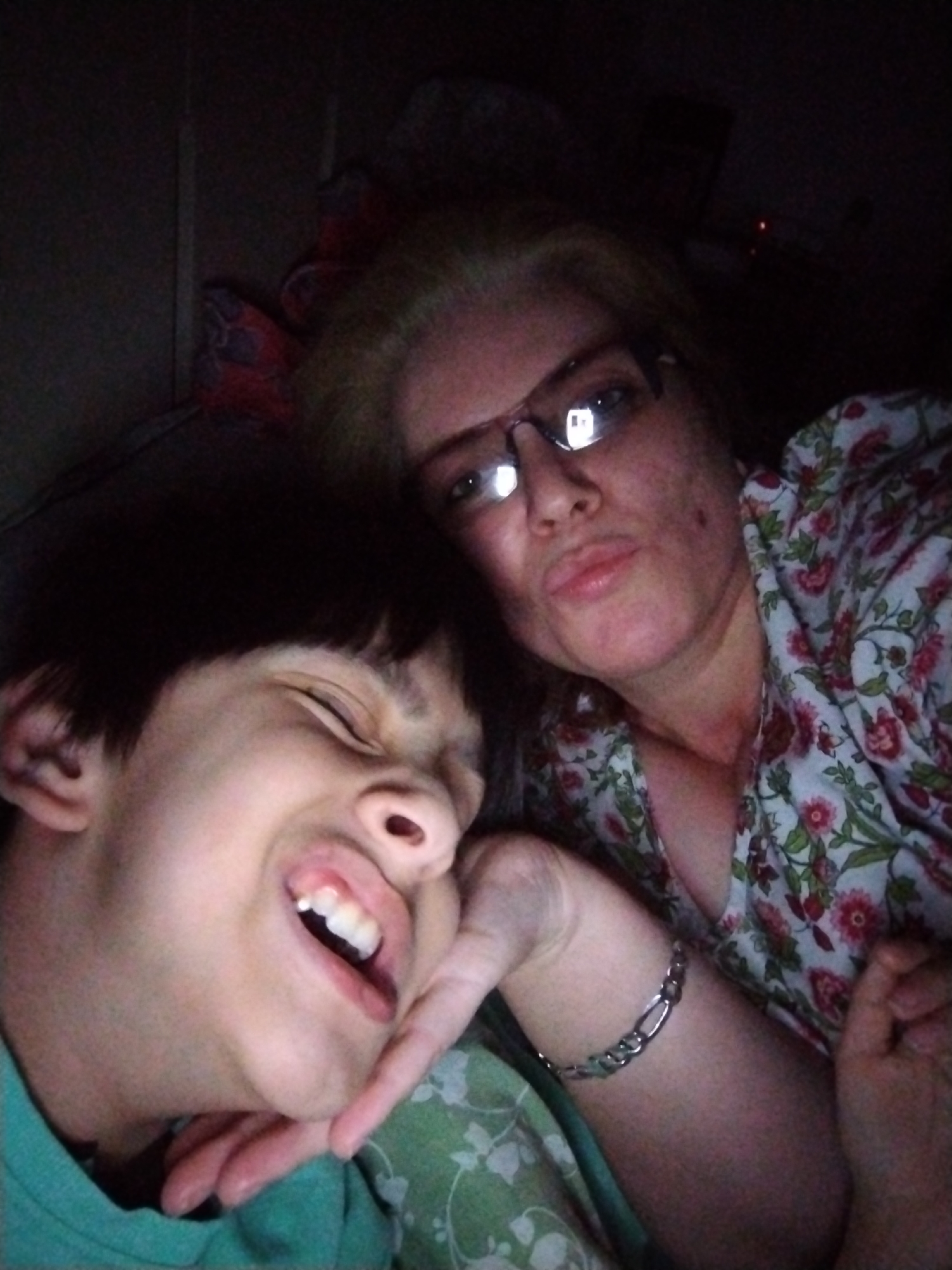 The image captures two individuals, an adult female and a younger person, in a close-up, dimly lit indoor setting. The precise geographical location, including Al Mas'oudi, United Arab Emirates, is not discernable from the visual evidence.
The adult female, positioned on the right, has light-colored hair and wears dark-rimmed eyeglasses that reflect a bright, rectangular light source, suggesting a screen. Her lips are pursed, directed towards the camera. Her skin exhibits multiple small, dark lesions or freckles across her face and neck. She is clad in a light-colored garment patterned with red and pink flowers and green leaves. A silver-toned, linked bracelet adorns her left wrist, and her left hand is positioned on the chin/jaw area of the younger individual.
The younger person, a child or adolescent with dark hair, is positioned on the left, facing away from the camera with eyes mostly closed and mouth open in a wide expression, revealing upper teeth, indicative of laughter or a playful grimace. They are wearing a teal or light green garment. Their right hand is partially visible, touching the palm of the adult female's hand.
The background is predominantly dark and indistinct, with limited details of a patterned fabric, potentially bedding, visible beneath the younger individual. The overall illumination is low, suggesting an ambient light source or light emanating from the device used to capture the image.
