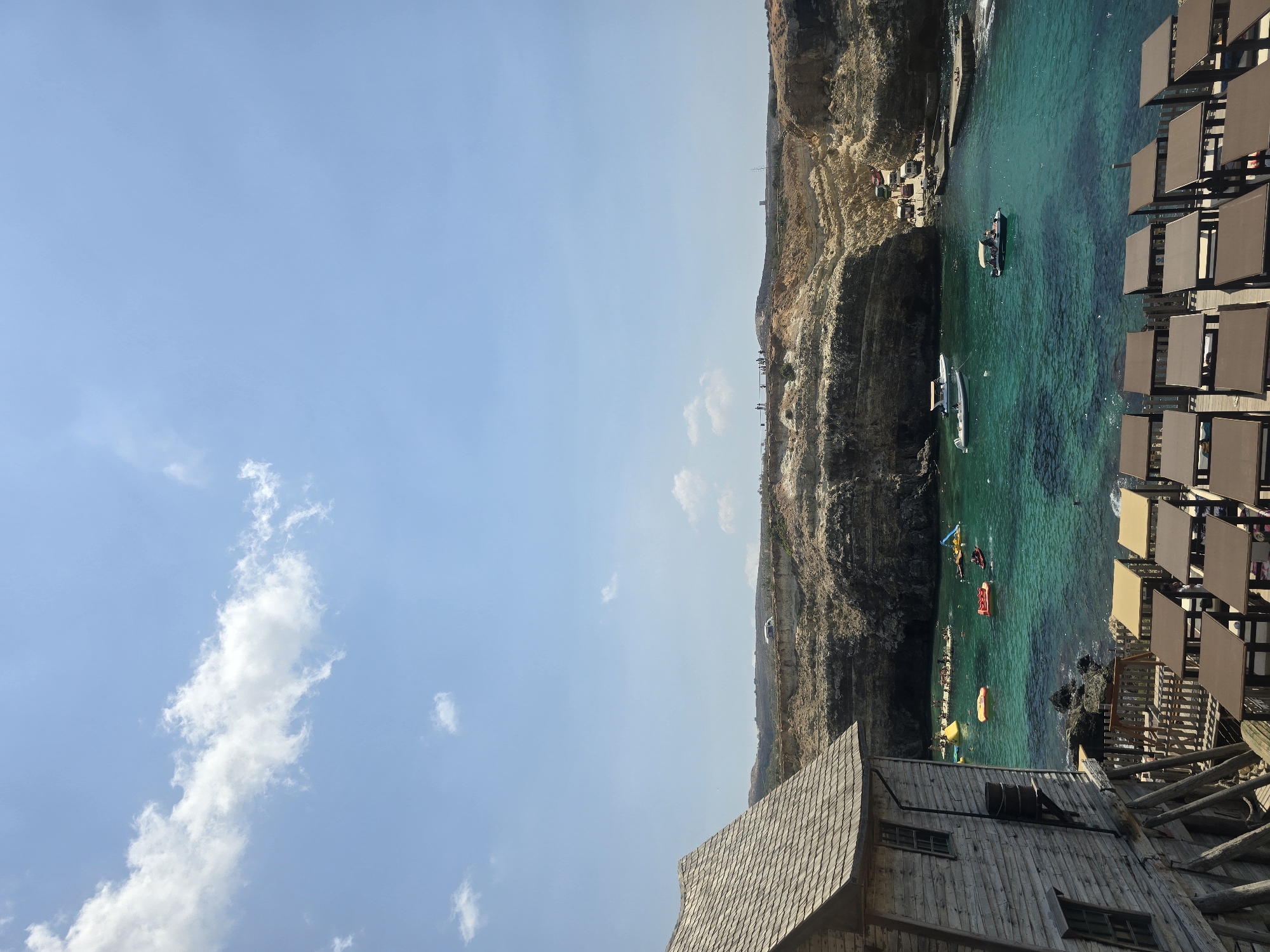 The image depicts a coastal scene in the city of Mellieħa, Malta, likely at or near Popeye Village, a popular tourist attraction. In the foreground, a rustic wooden building with a shingled roof and visible wooden planks stands to the left. Adjacent to it, a series of outdoor tables are set up, some with brown tabletops and others with tan. A wooden fence encloses a small area with what appears to be a swing set.
The middle ground reveals a vibrant turquoise bay filled with activity. Numerous inflatable rafts and small watercraft, including a yellow raft, a red raft, and a yellow banana boat, are scattered across the water. Several people are visible on and around these inflatables, enjoying the water. Two motorboats are also present, one white with an orange canopy and another smaller white boat with a beige canopy, carrying passengers. The bay is bordered by dramatic, rugged cliffs with layered rock formations, suggesting a natural cove or inlet. Small rocks are visible along the shoreline.
In the background, the cliffs continue, with a pathway or road carved into the cliffside. Several vehicles, including what appears to be a white van and other cars, are parked near a concrete jetty or ramp on the right side of the bay. A few people are visible near the vehicles and on the cliff edge, observing the scene.
The sky above is a clear, bright blue with scattered white, fluffy clouds. The overall impression is a sunny, lively day at a popular coastal tourist destination in Malta.