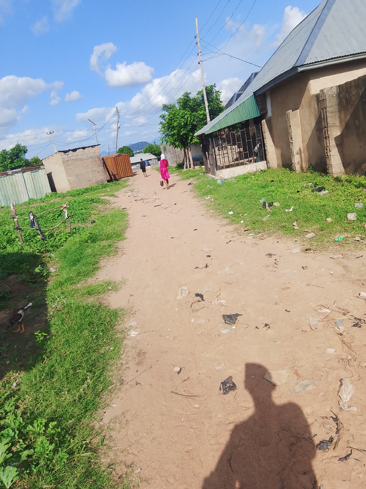 This is a ground-level, eye-level photograph of a dirt road in the city of Jalingo, Nigeria. The road is lined with a mix of buildings, vegetation, and some scattered debris. On the right side of the road, a building with a corrugated metal roof and concrete walls is prominent. The building appears to be a residential or commercial structure, with a green awning over an open area with a metal grate. To the left of the road, there are some smaller, unfinished concrete block structures and a metal fence. Lush green grass and some low-lying plants grow along the sides of the road. Further down the road, two individuals are walking away from the camera. One is wearing a bright pink dress and the other is wearing dark clothing. A chicken is visible in the lower left portion of the image, pecking at the ground. The sky is blue with scattered white clouds. The overall impression is of a rural or semi-urban environment in Jalingo, Nigeria, likely a neighborhood with a mix of completed and under-construction dwellings.